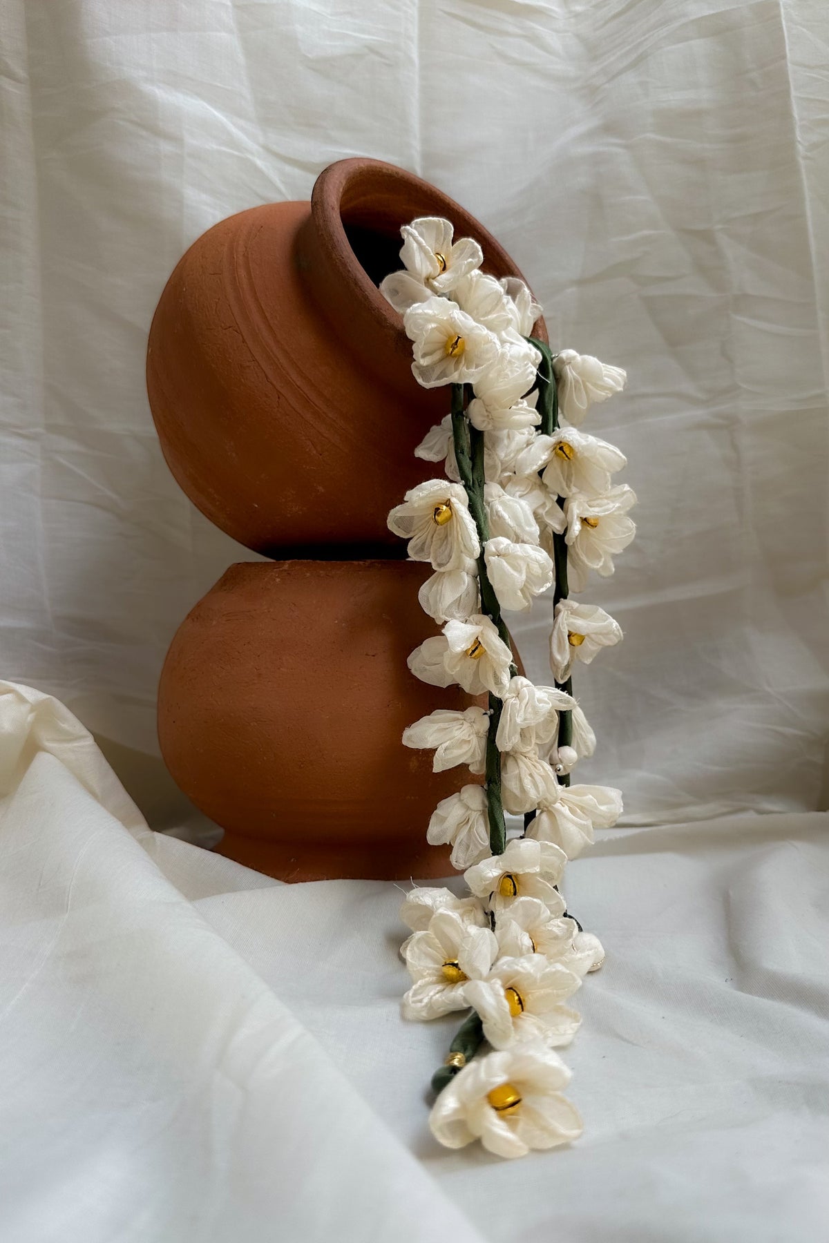 White handmade flower garlands draped over a stacked terracotta ceramic vase against a draped white fabric background.