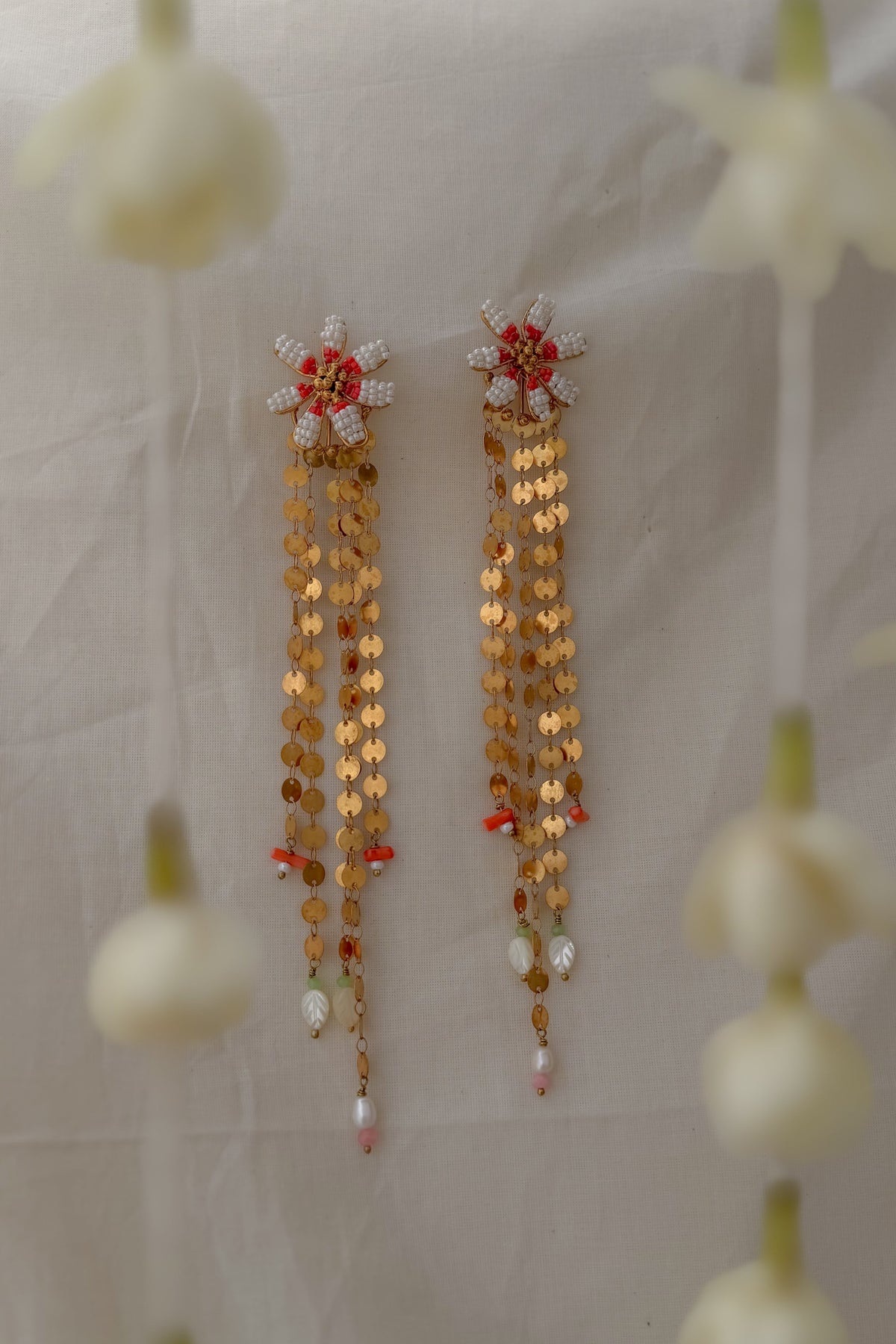 A pair of traditional Indian gold-toned dangle earrings with red and white floral motifs, hanging against a draped off-white fabric backdrop with blurred white flower decorations in the foreground.