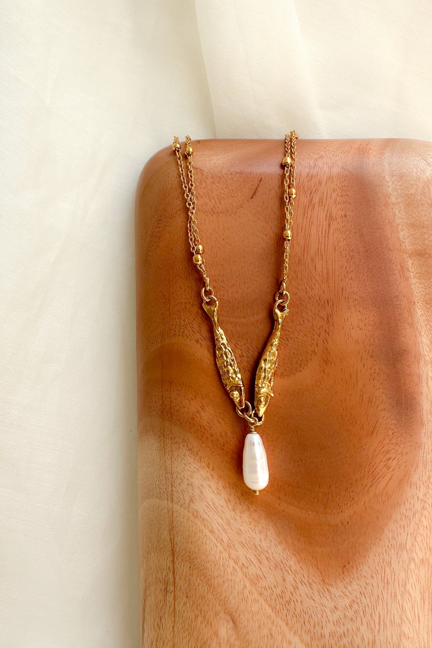 Fish charm Gold necklace with a white Pearl pendant on a wooden block against a neutral background