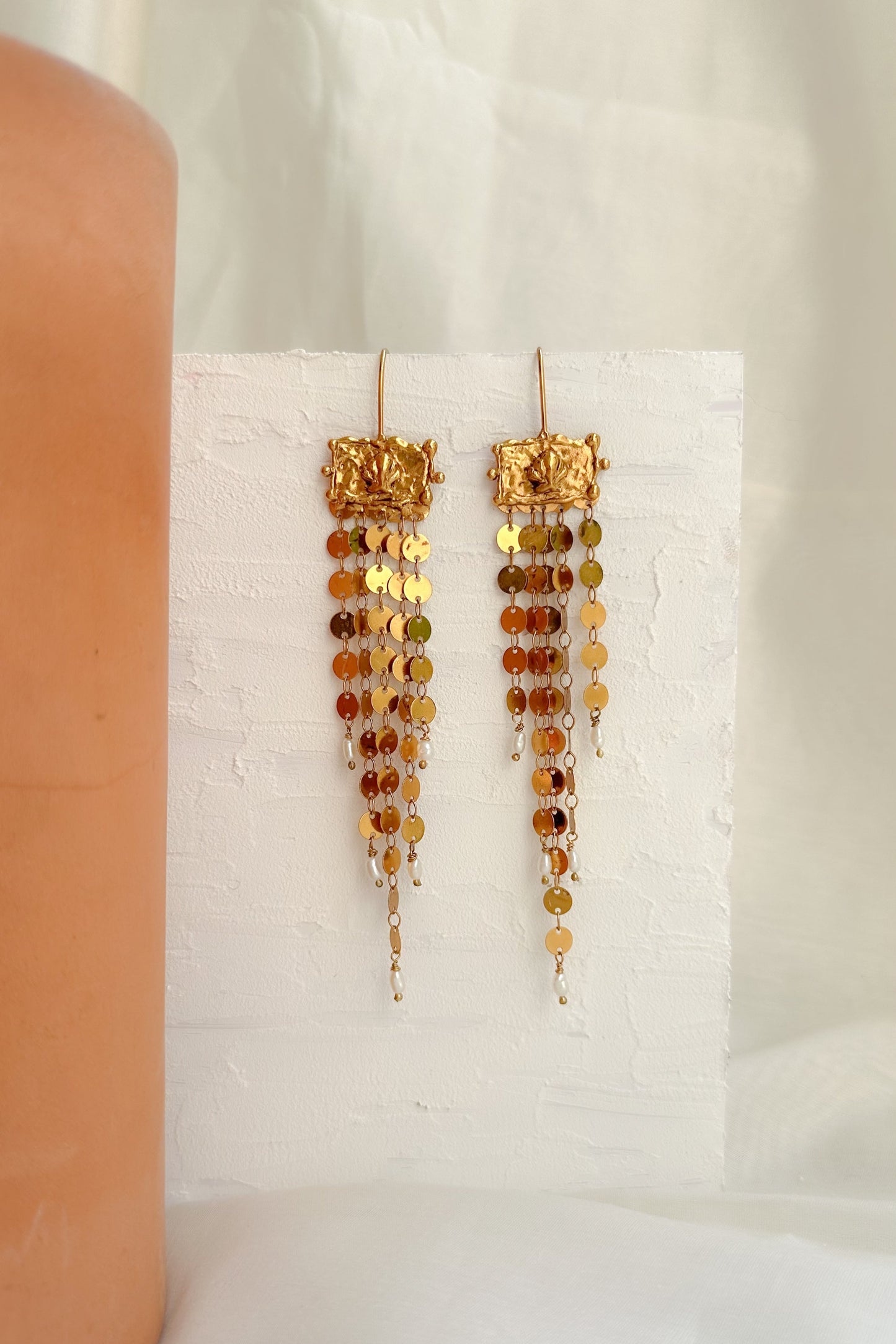 Pair of gold and amber beaded earrings on a white stand with a beige candle in the background.