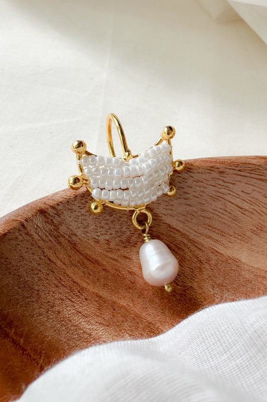 A gold moon ear cuff / bugadi with a white pearl-encrusted crown-like top and a dangling teardrop pearl, displayed on a wooden surface with white fabric.