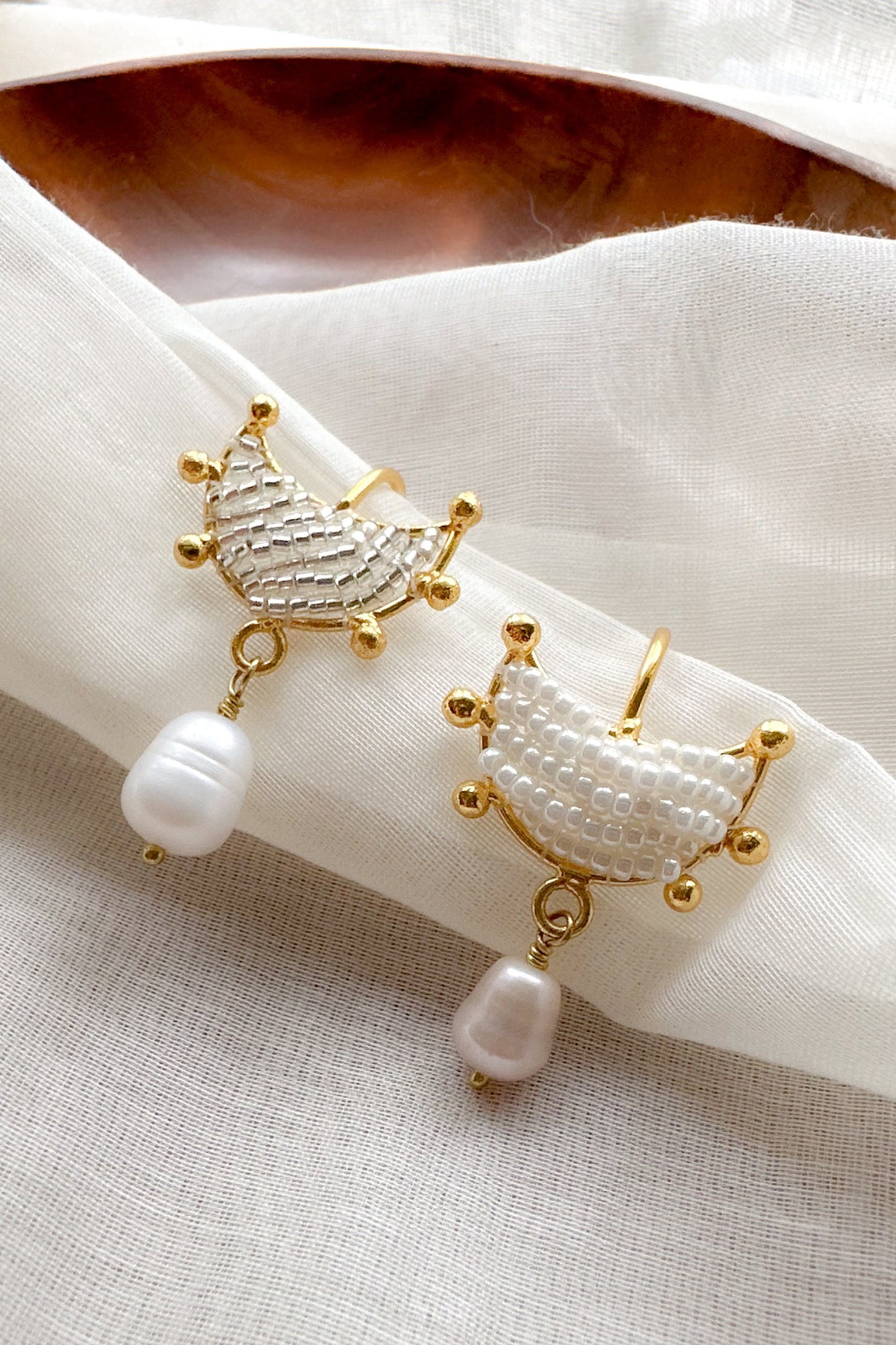 A pair of gold crescent-shaped dangle ear cuffs with textured white beadwork and a dangling white pearl, displayed on a white fabric napkin.