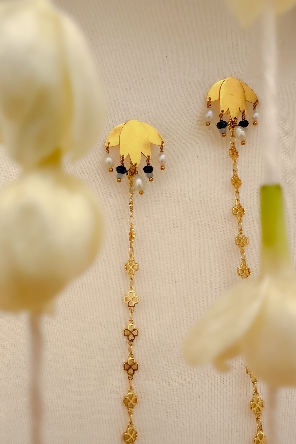A pair of gold-toned traditional Indian earrings with floral tops, small white pearl-like beads, and long dangling chain elements, displayed against a soft cream background with blurred white flowers.