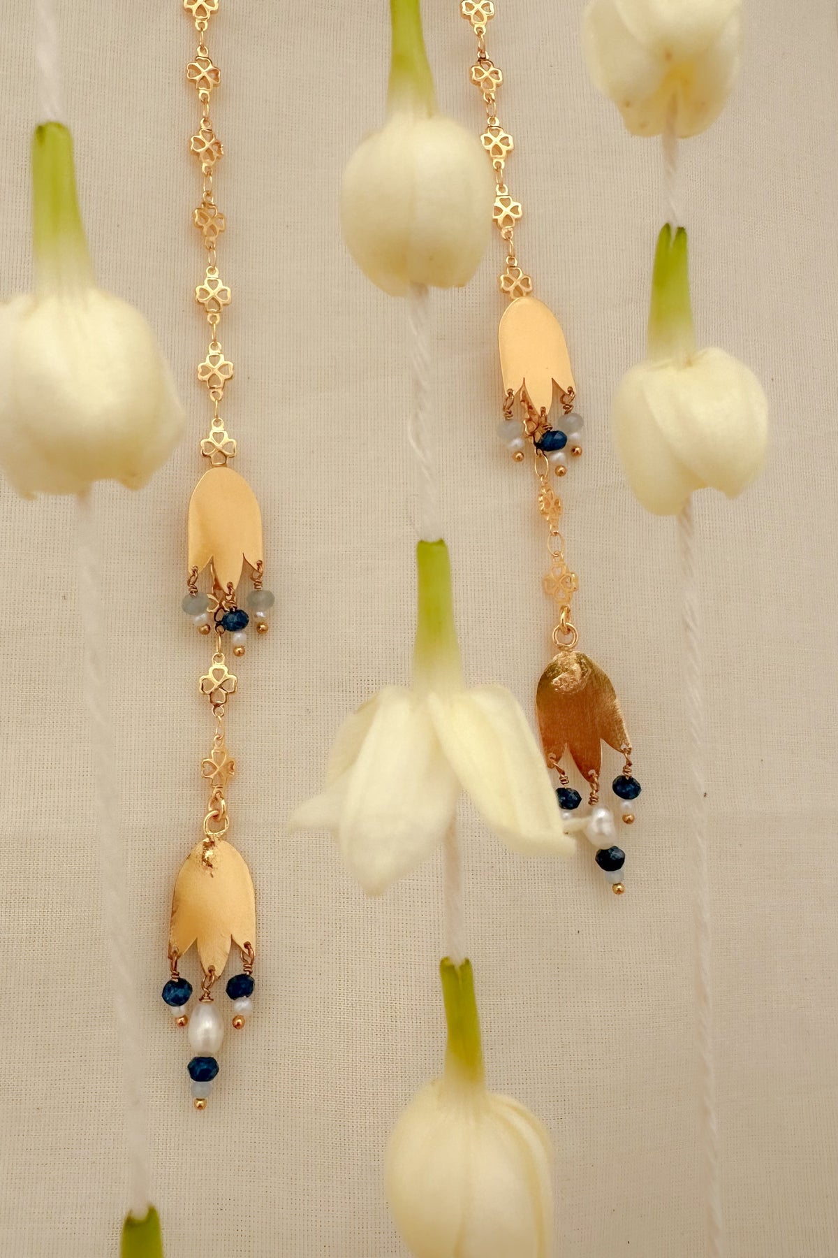 Gold-toned floral dangle earrings with cream flower petals, small blue beads, and pearl  accents, shown hanging against a light neutral background with white flower props.