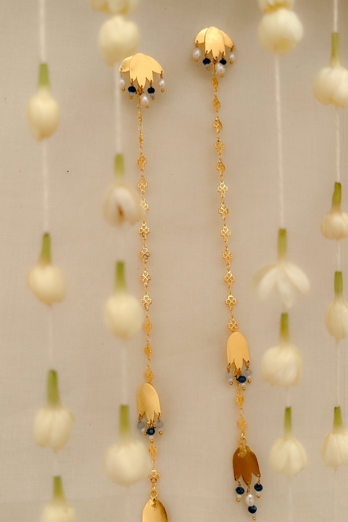A pair of gold-toned traditional Indian earrings with hanging floral elements, small white pearls, and dark blue stones, displayed against a soft beige background with hanging white flowers.