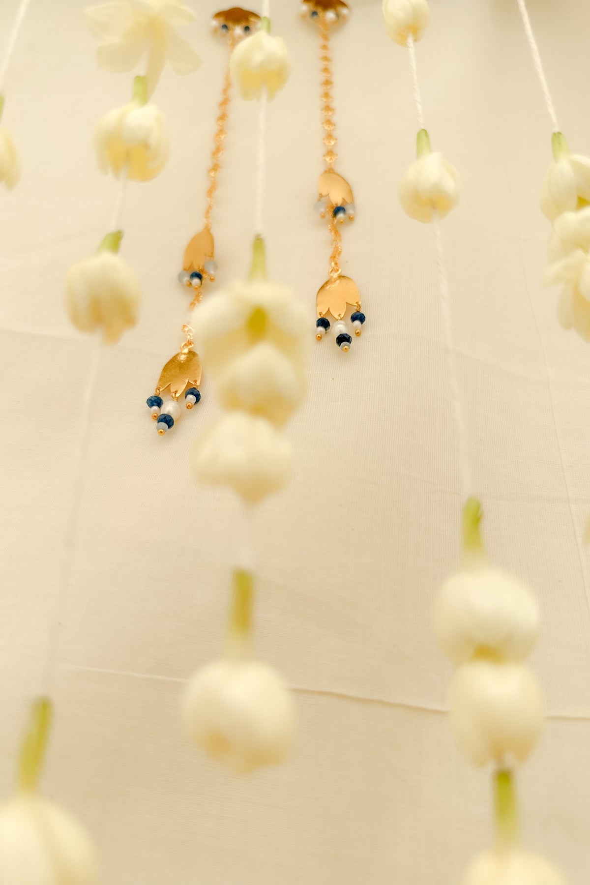 Gold-toned dangling earrings with small blue bead accents, hanging against a soft cream fabric backdrop with blurred white flower-like decorations.
