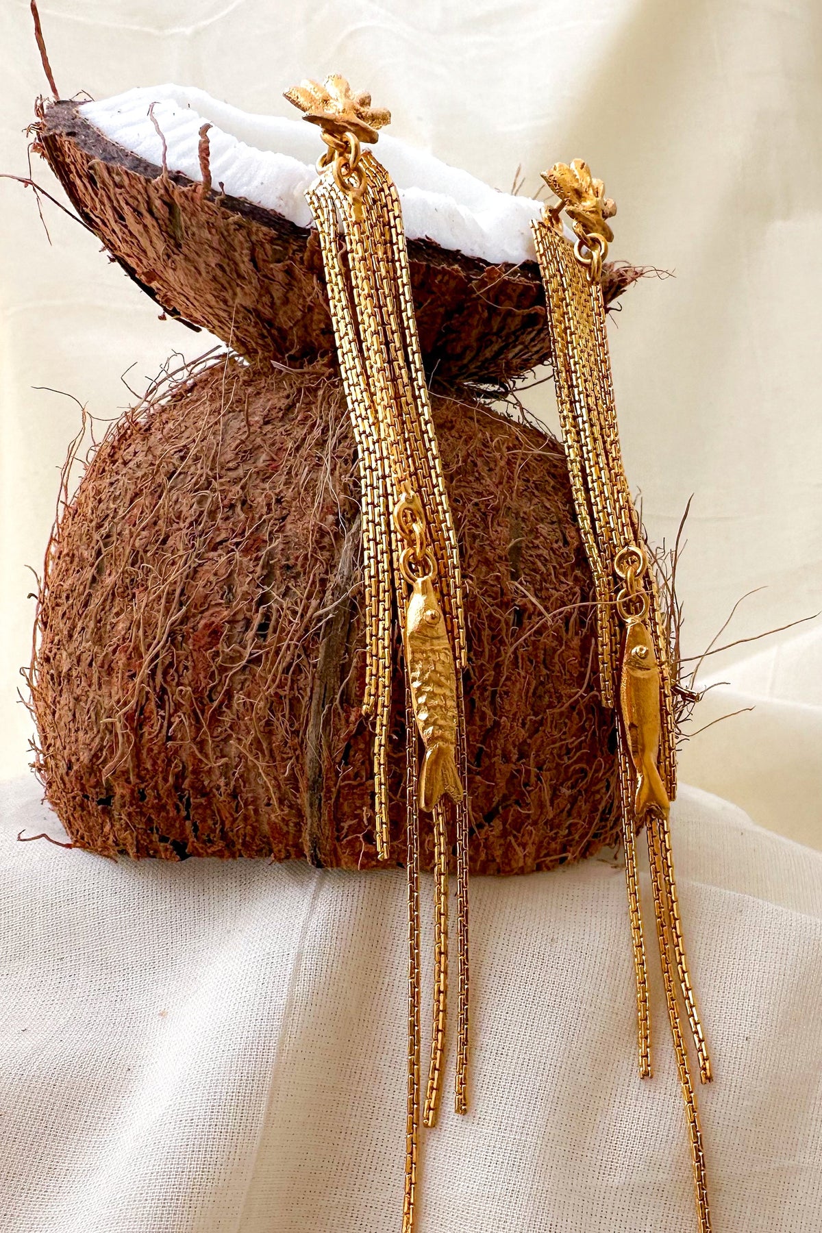 A pair of gold-toned traditional Indian earrings with fish-shaped charms and dangling chain tassels, displayed on a coconut shell prop against a draped cream fabric background.