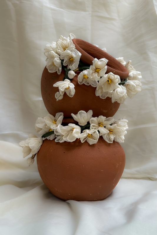 A terracotta-colored ceramic vase with a rounded, two-tiered design, decorated with white artificial flowers with yellow centers, set against a draped white fabric background.