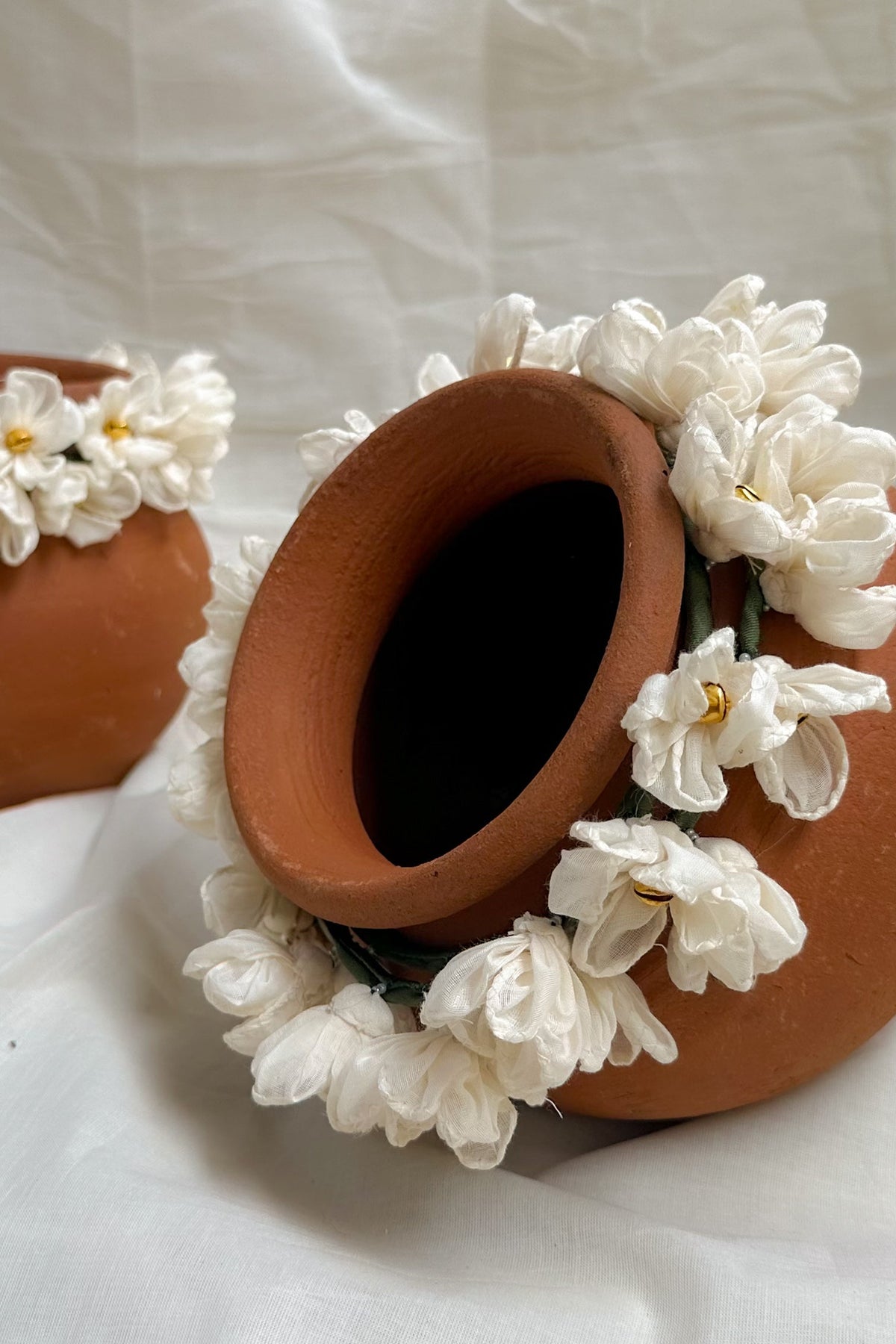 Two terracotta clay pots decorated with white flowers, arranged on draped white fabric.