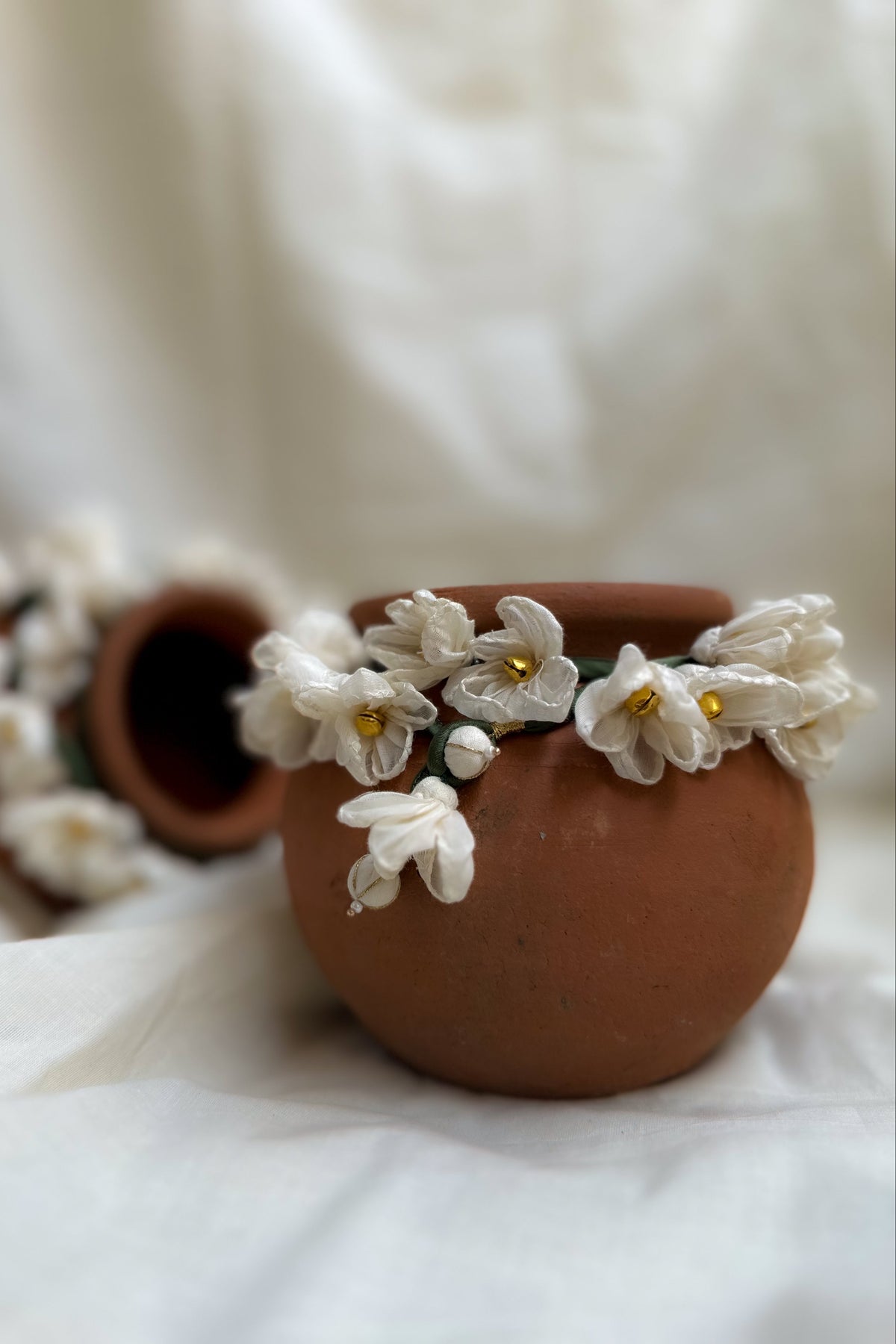 A terracotta clay pot decorated with white fabric flowers and small gold centers, shown on draped white fabric with another similar pot blurred in the background.