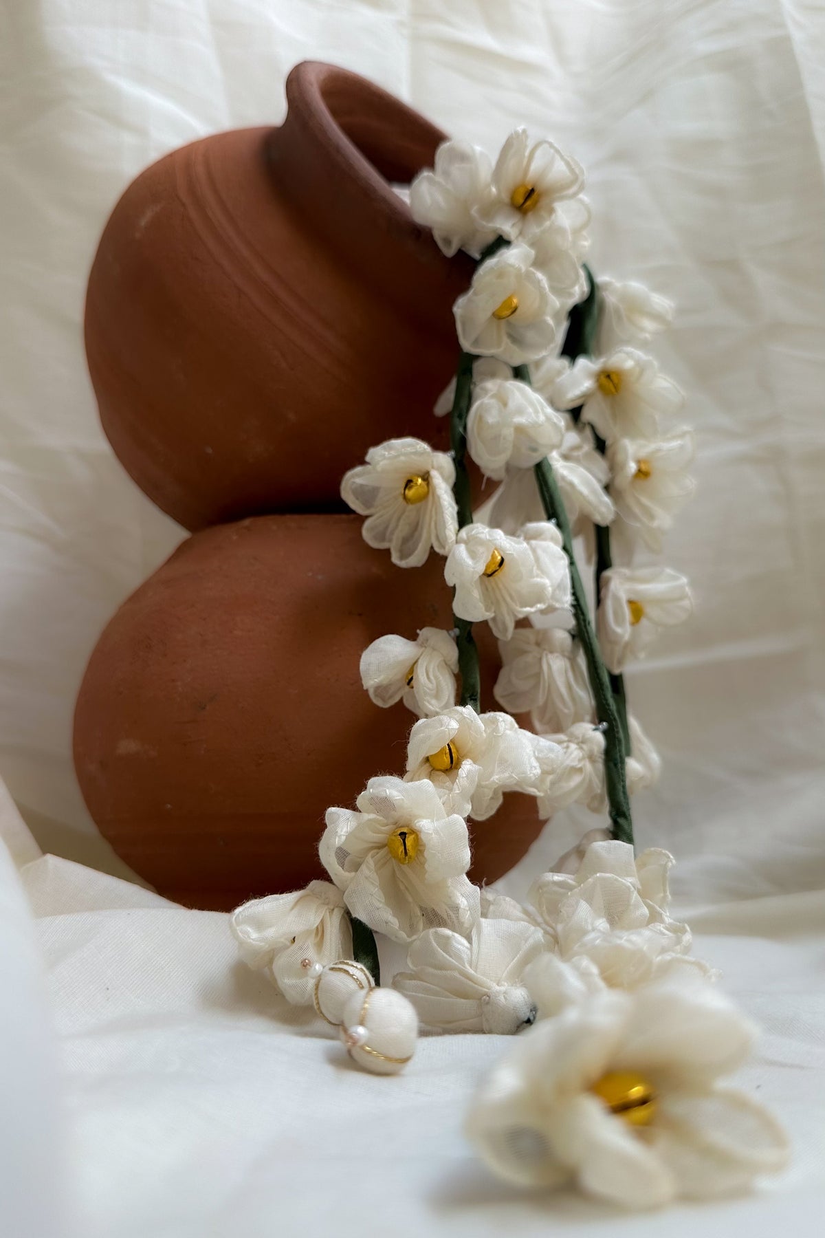 White artificial daffodil garlands with yellow centers draped over stacked terracotta pots on a white fabric background.