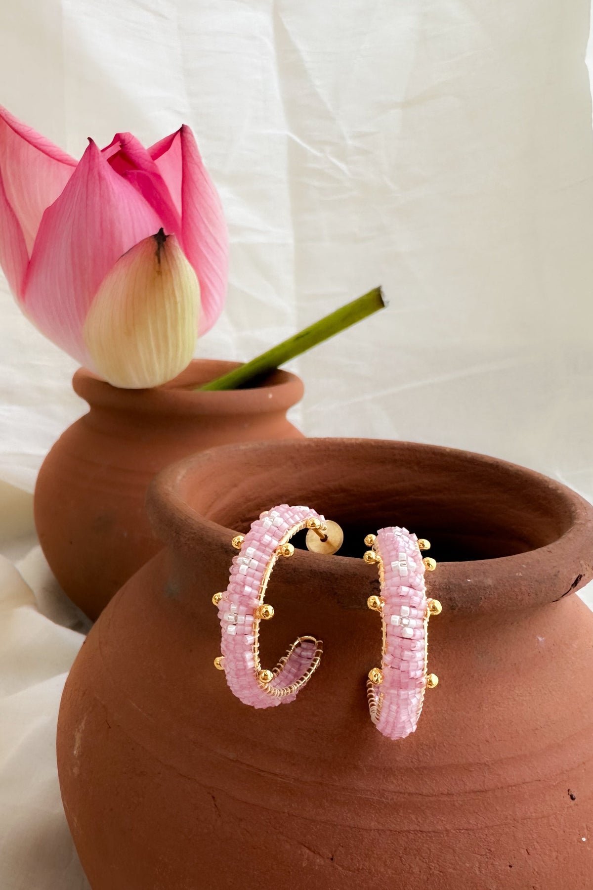 Pink beaded hoop earrings with gold-toned studs, draped over a terracotta pot with a pink lotus flower in the background.