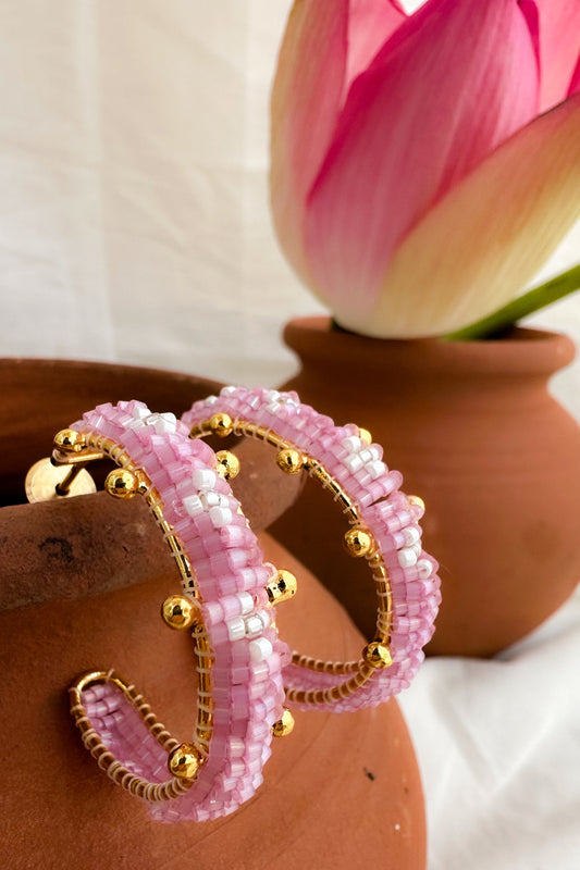 A pair of pink beaded hoop earrings with gold-toned metal wire detailing and small gold beads, displayed on a terracotta pot with a pink lotus flower in the background.