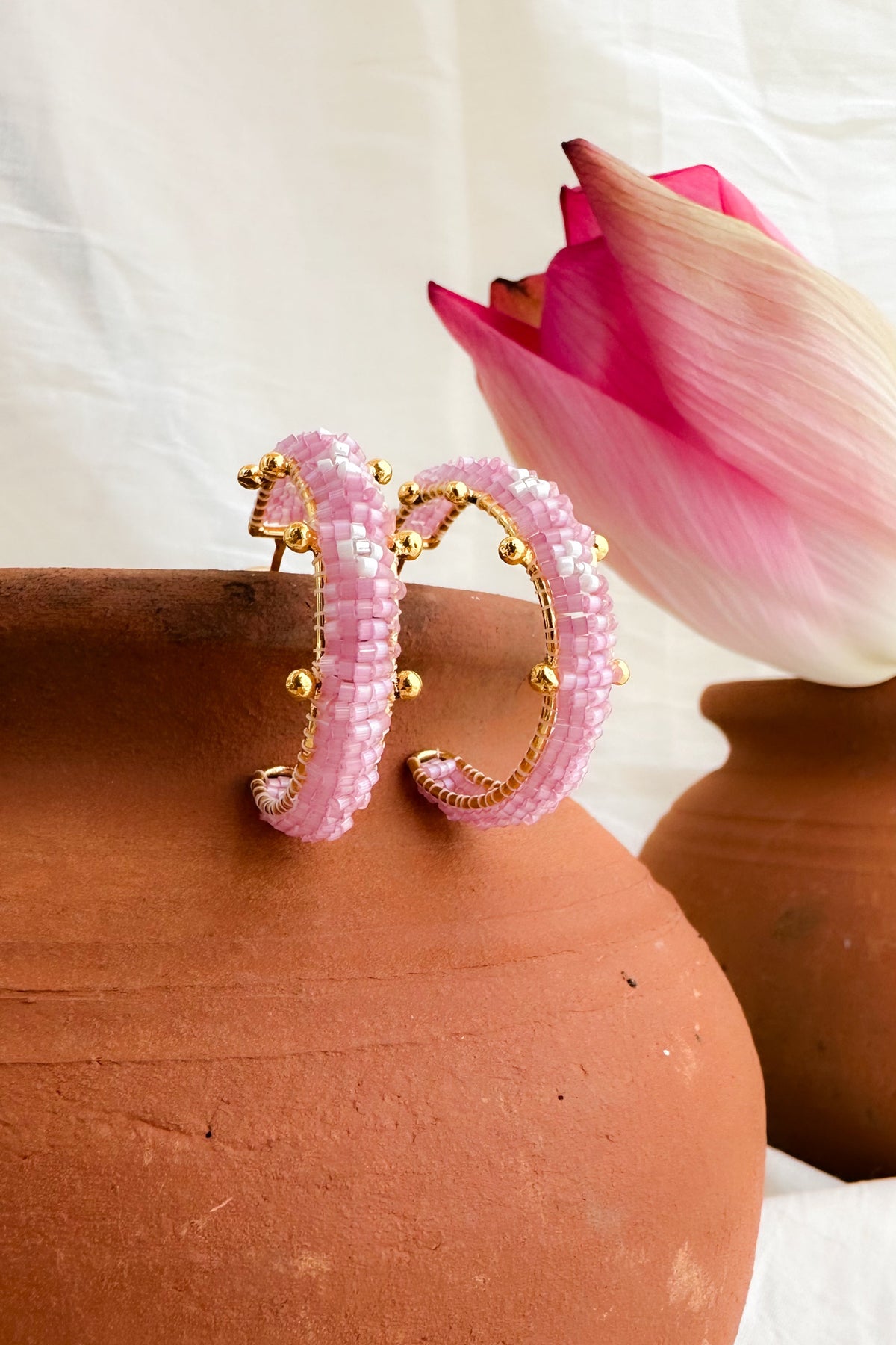 A pair of pink beaded hoop earrings with gold-toned metal trim and small gold bead accents, displayed on a terracotta pot with a pink lotus flower in the background.