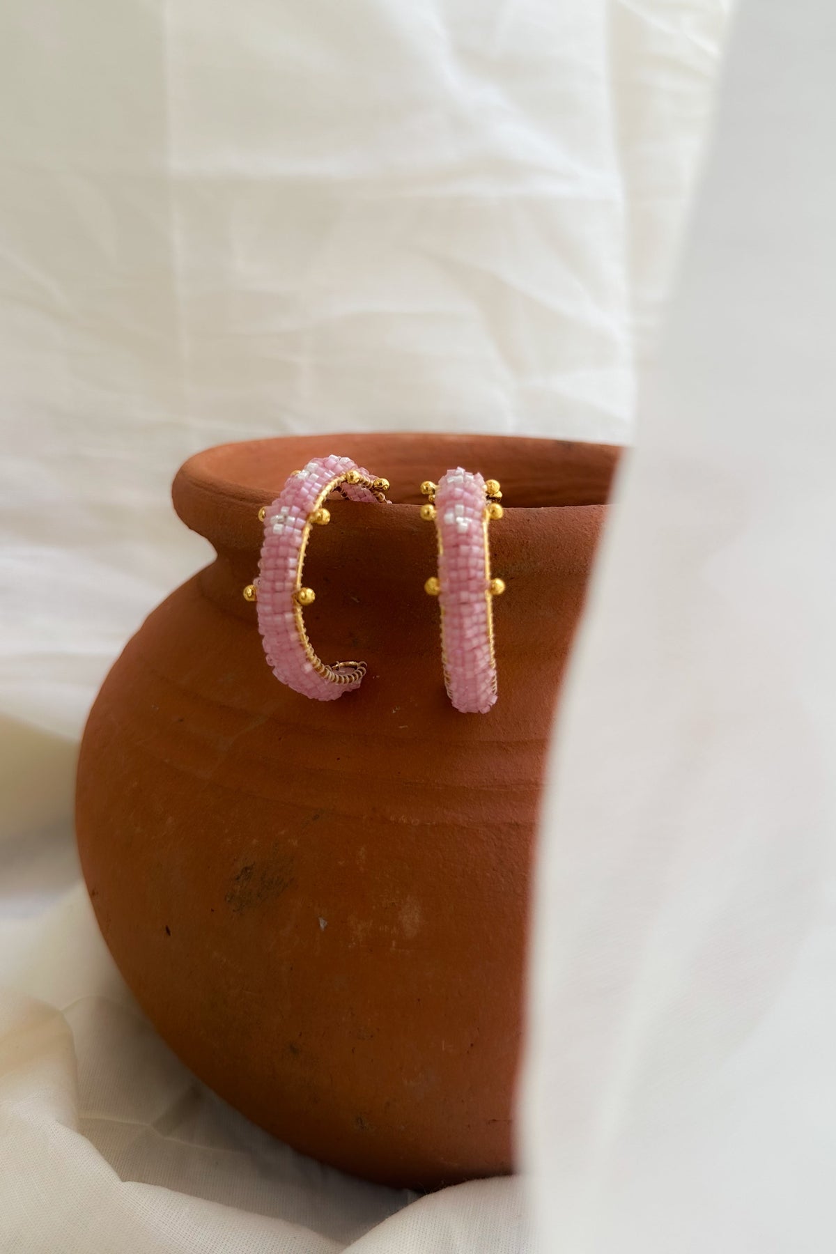 A pair of gold hoop earrings with textured pink inlay, resting on a terracotta pot against a draped white fabric background.