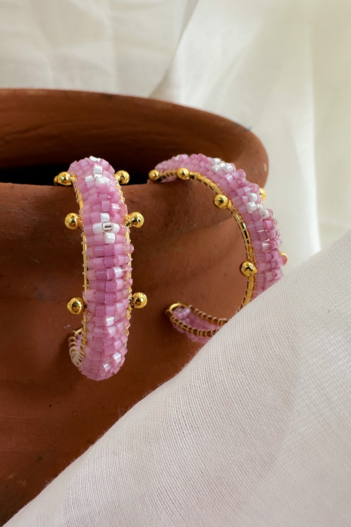 A pair of pink beaded huggie earrings with gold-toned metal studs, displayed on a brown ceramic surface with a white fabric background.