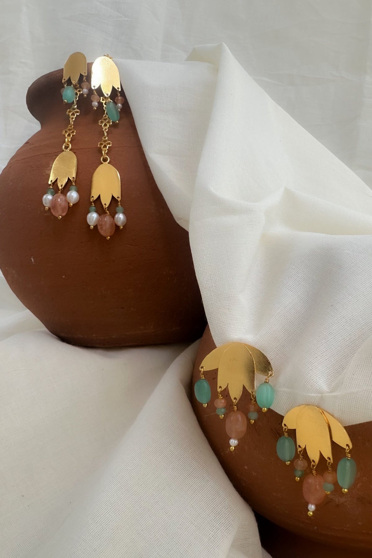 Gold-toned statement earrings with layered petal-shaped charms and dangling turquoise, pink, and pearl beads, displayed draped over a terracotta pot on a white fabric backdrop.