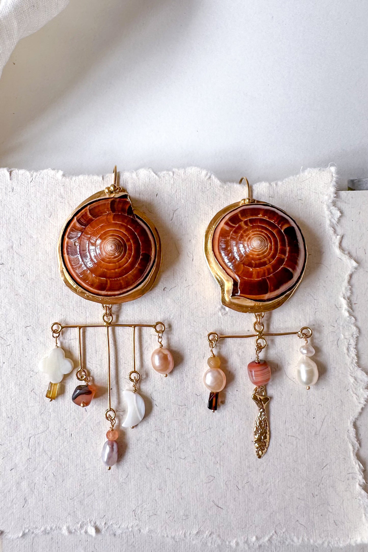 Pair of ornate earrings with shell-like designs and pearl beads on a textured white surface.