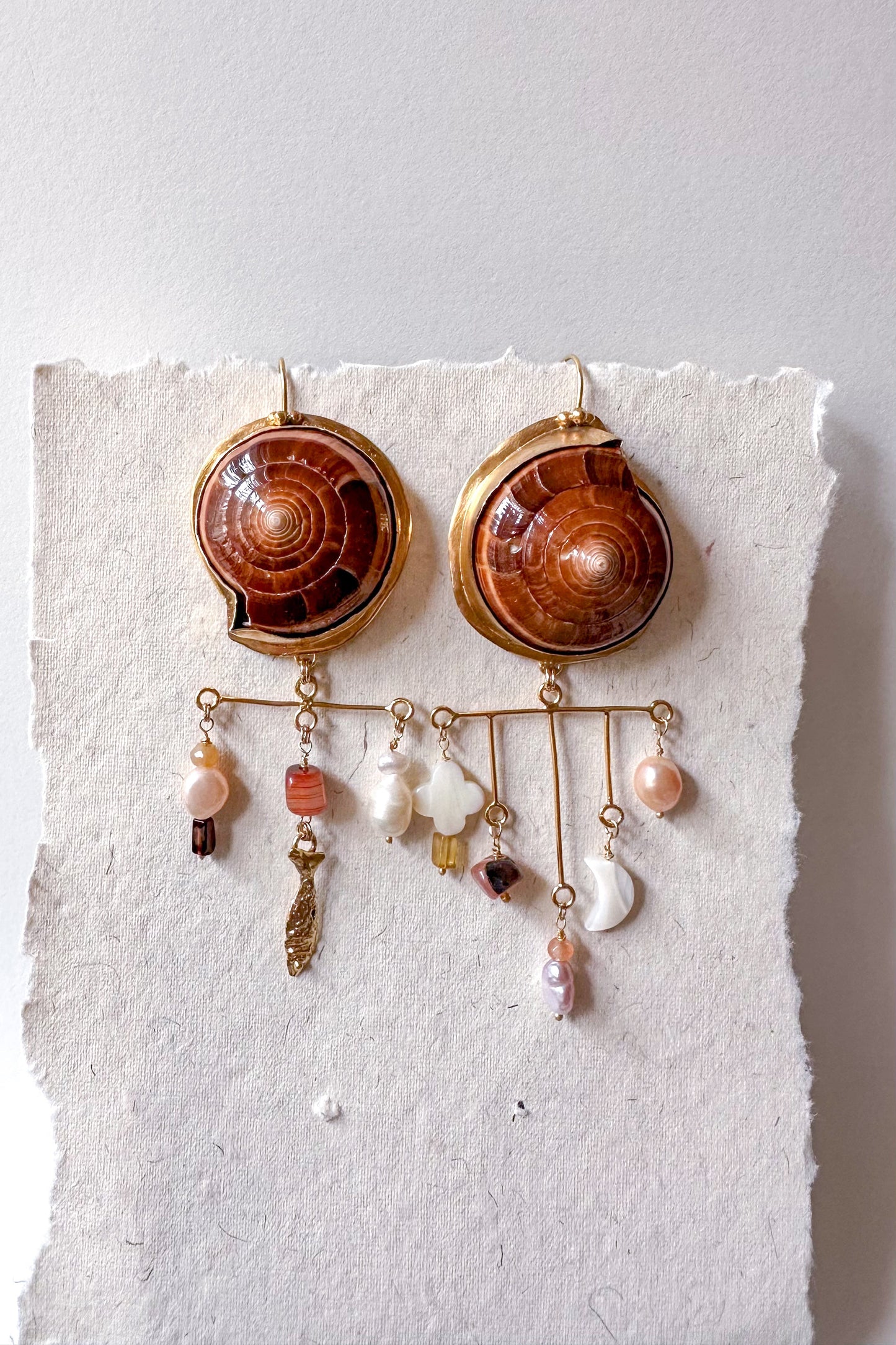 Pair of earrings with shell-like designs and hanging charms on a textured white surface.