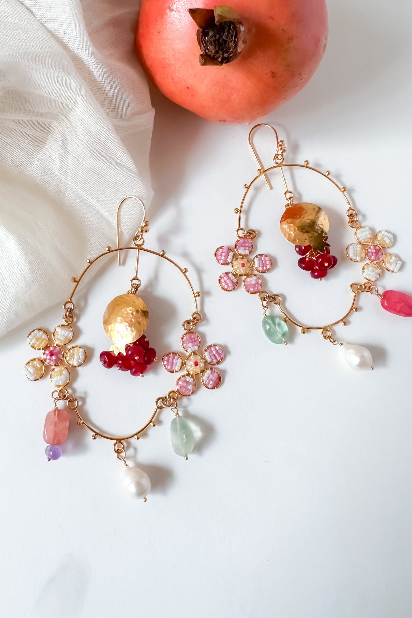 Colorful hoop earrings with gemstones and pearls next to a pomegranate on a white fabric background.