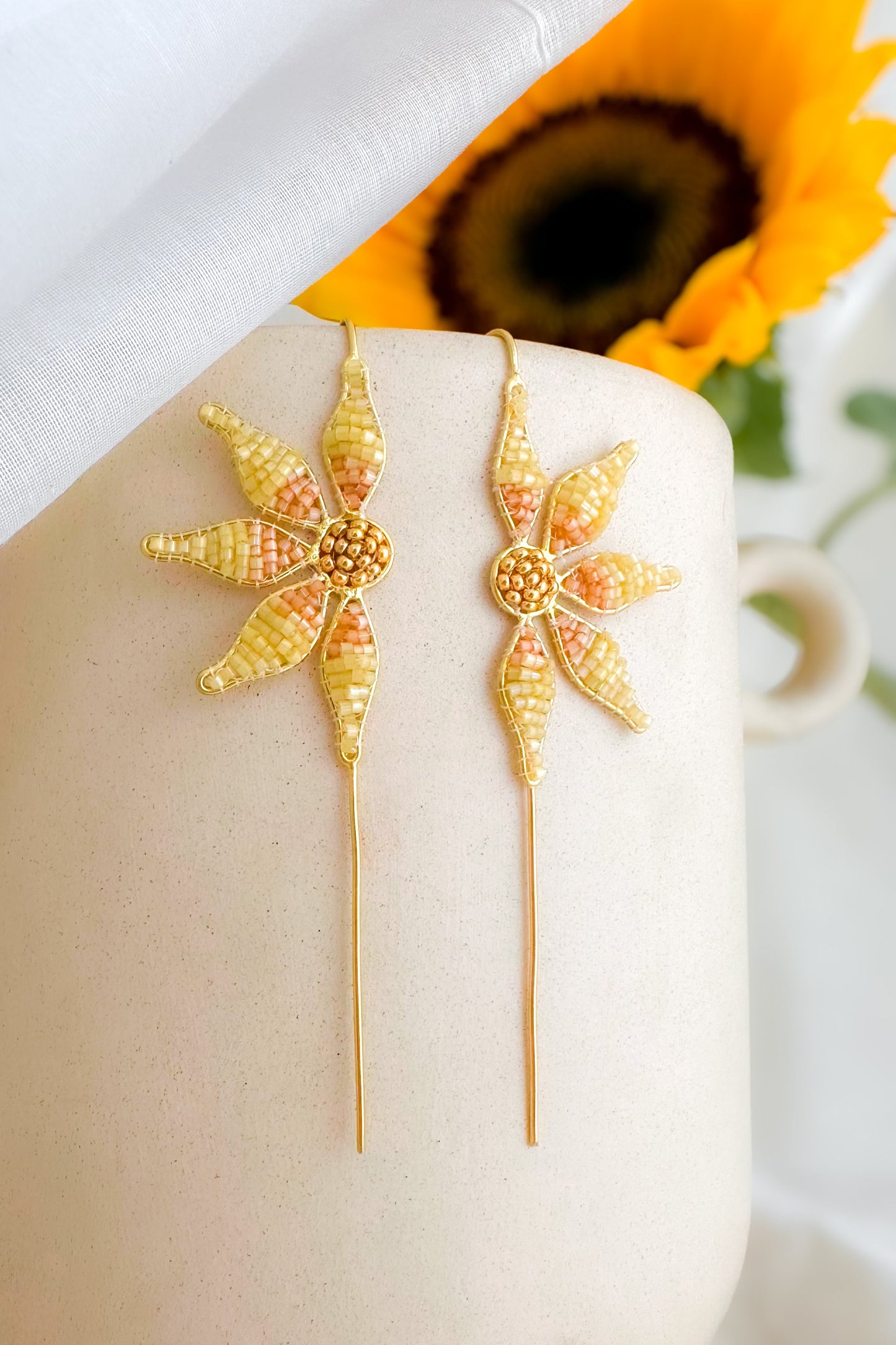 Gold floral earrings on a white surface with a sunflower in the background