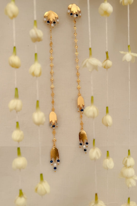 A pair of long gold-toned traditional earrings with floral motifs and small dark blue bead accents, hanging against a light neutral background with decorative white flowers.