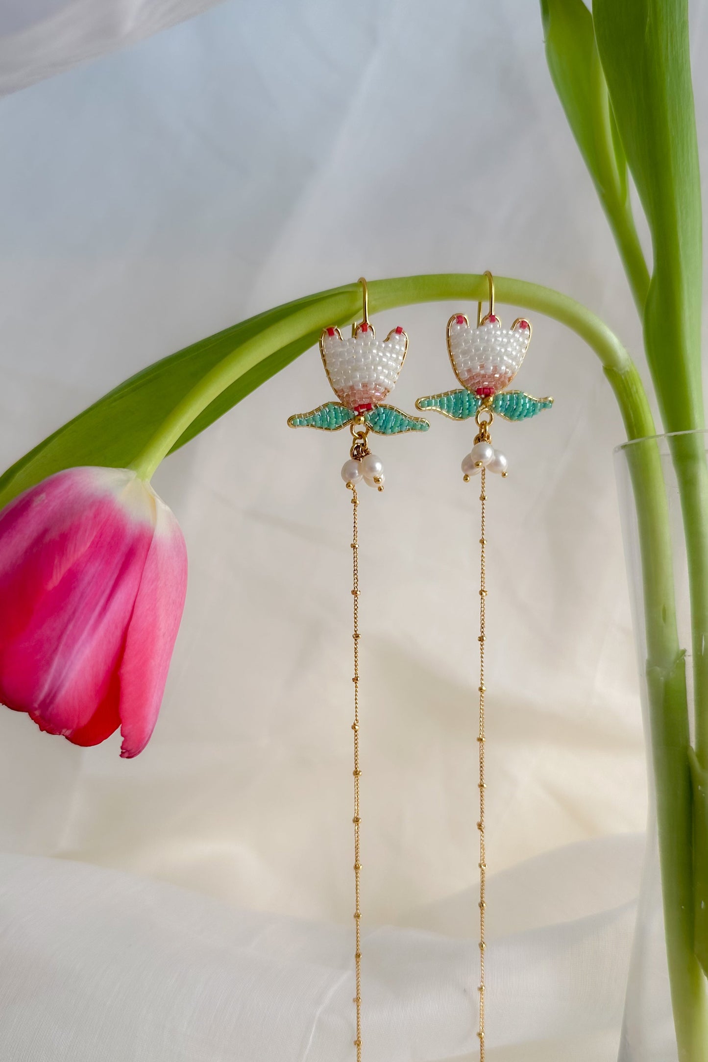 Earrings with floral design hanging from a pink tulip against a light background