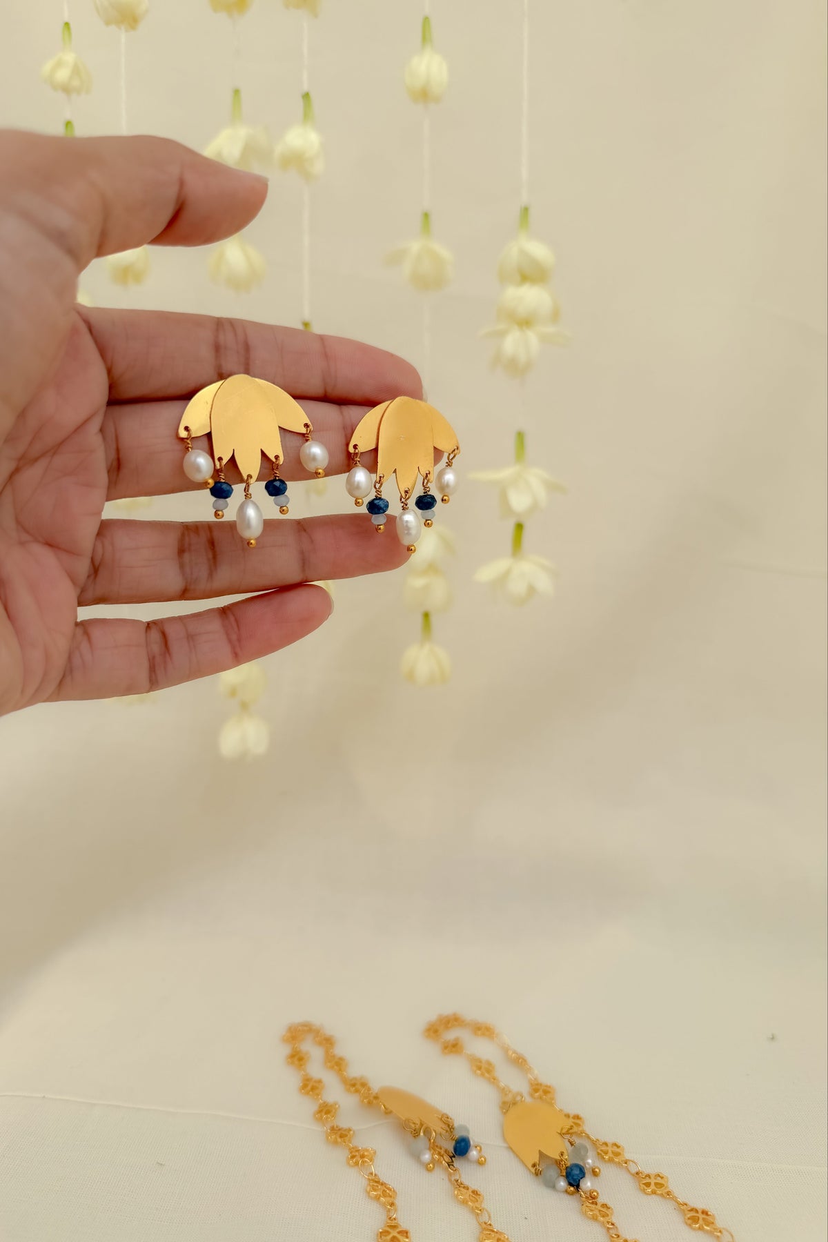 A hand holds a pair of gold leaf-shaped earrings with white pearls and dark blue beads, with matching gold necklaces visible at the bottom against a cream background with hanging white flowers.