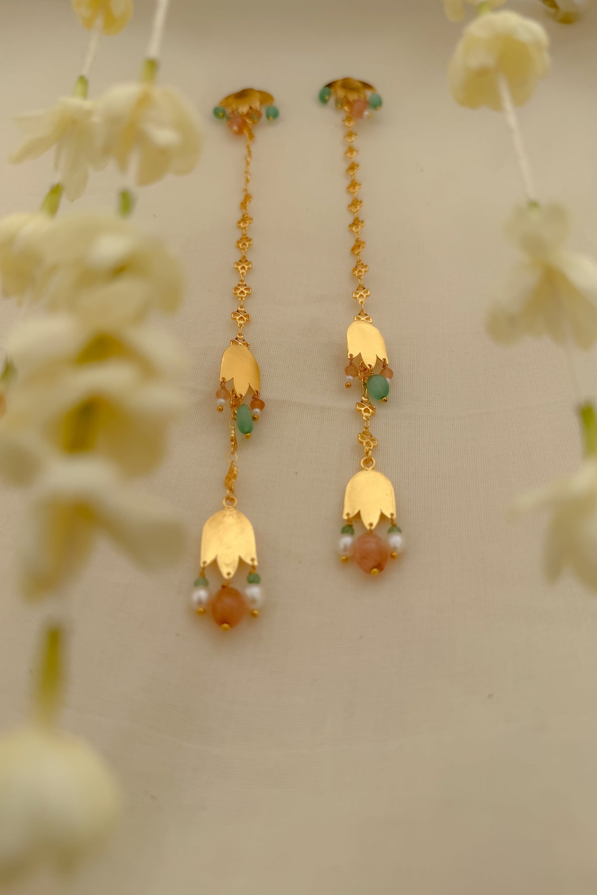 A pair of gold-toned traditional dangle earrings with green and peach stones and pearl-like accents, displayed against a light beige background with soft white flowers in the foreground.