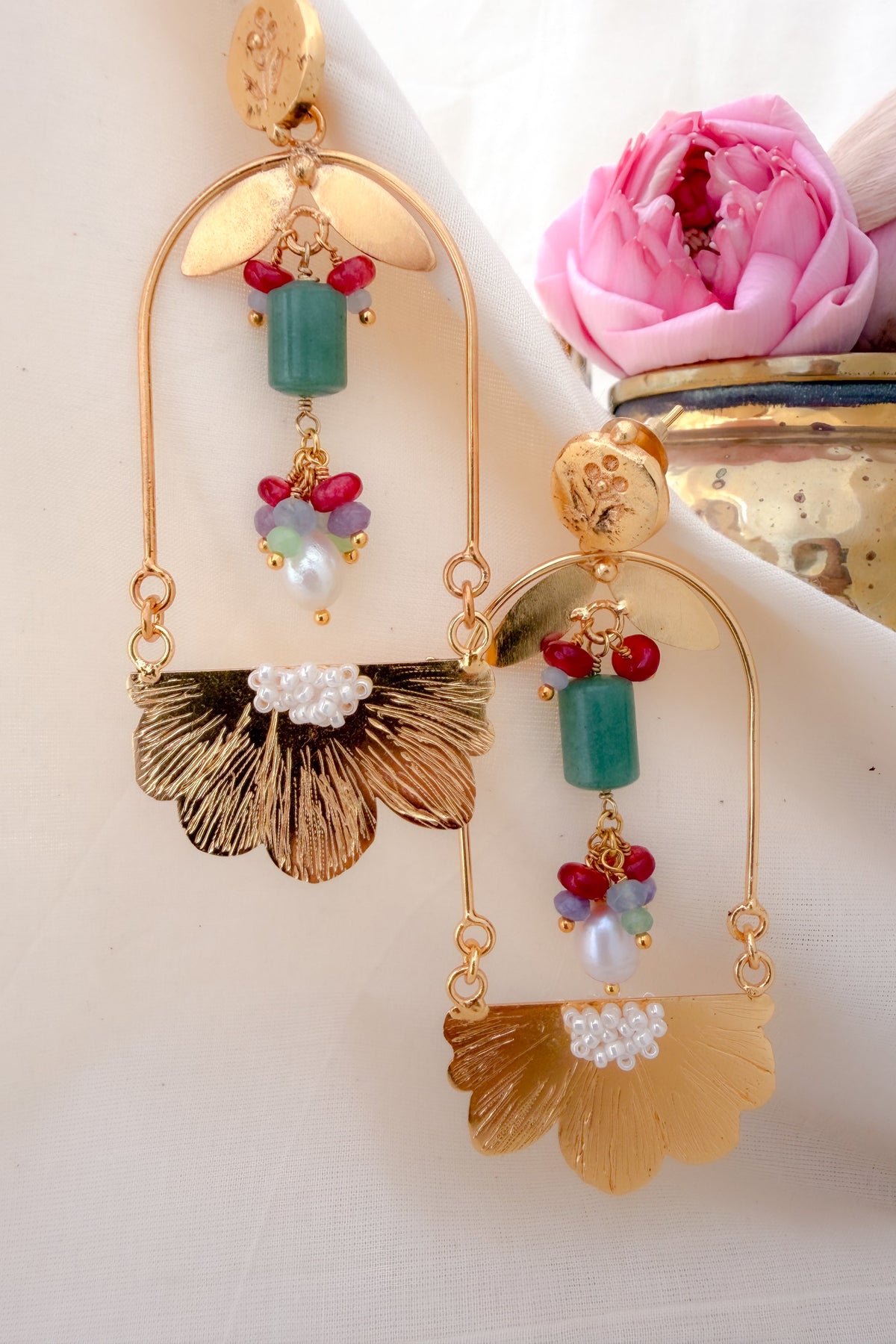 A pair of ornate gold-toned statement earrings with green cylindrical beads, red and lavender heart-shaped beads, and textured fan-shaped gold dangles, displayed on a white fabric background next to a pink flower.