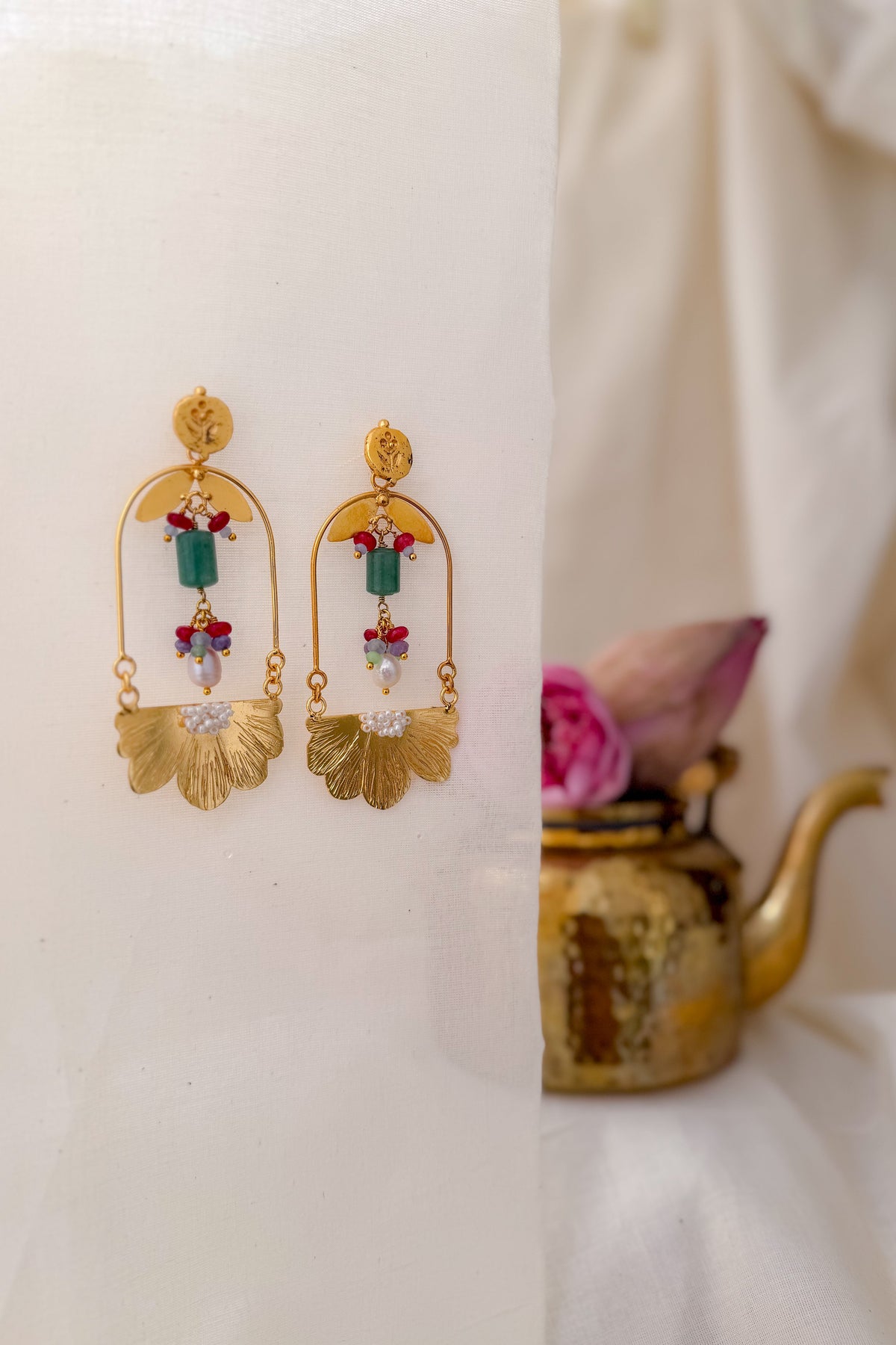 A pair of gold dangle earrings with green, red, and clear crystal details, displayed on a white draped fabric next to a small brass teapot with a pink flower.
