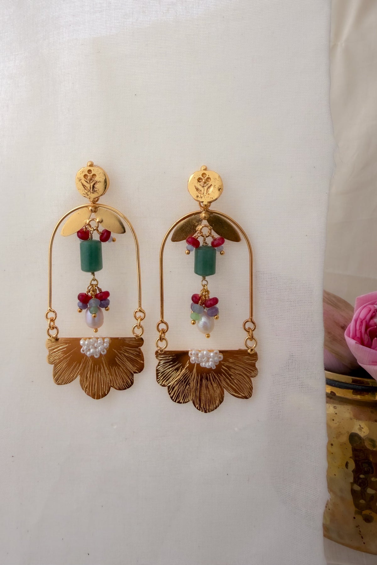 A pair of gold-toned statement dangle earrings with green rectangular beads, small red and blue beads, and white pearl-like accents, displayed against a white fabric backdrop.