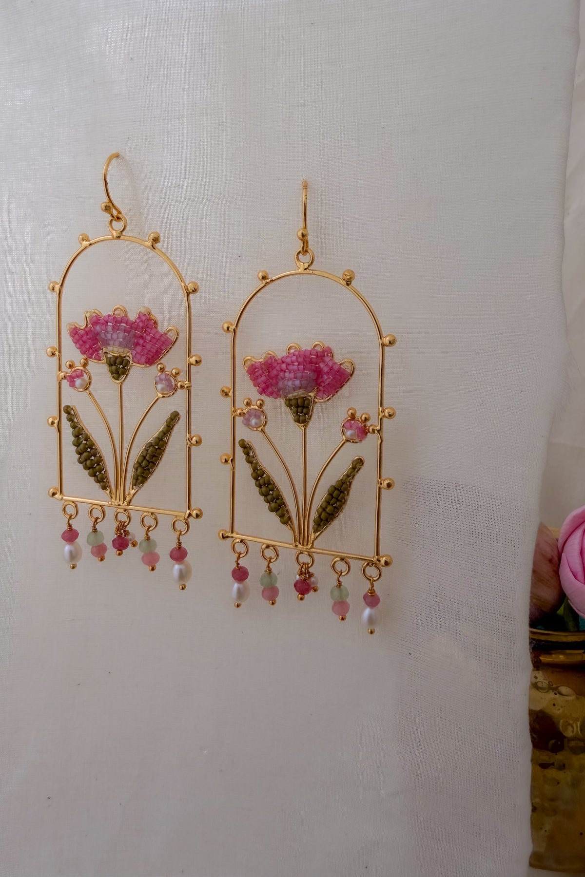 A pair of gold-toned dangle earrings with pink floral motifs, green leaf details, and dangling bead accents, displayed against a white fabric background.