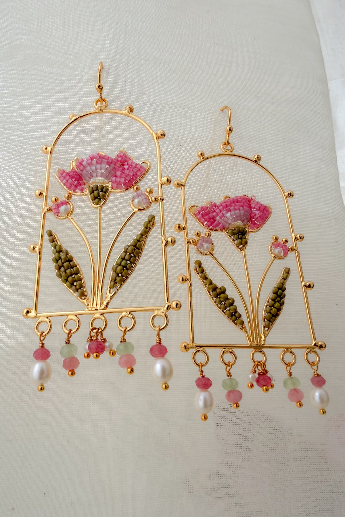 A pair of gold-toned traditional Indian earrings featuring pink and green beaded floral motifs with dangling pearl-like beads, displayed on a light fabric background.