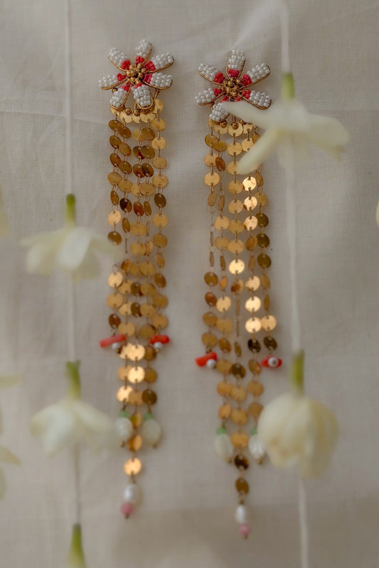 A pair of traditional Indian gold-toned sequin dangle earrings with white crystal-embellished flower motifs at the top and small red and pink bead accents, shown hanging against a light fabric backdrop with blurred white flowers around them.