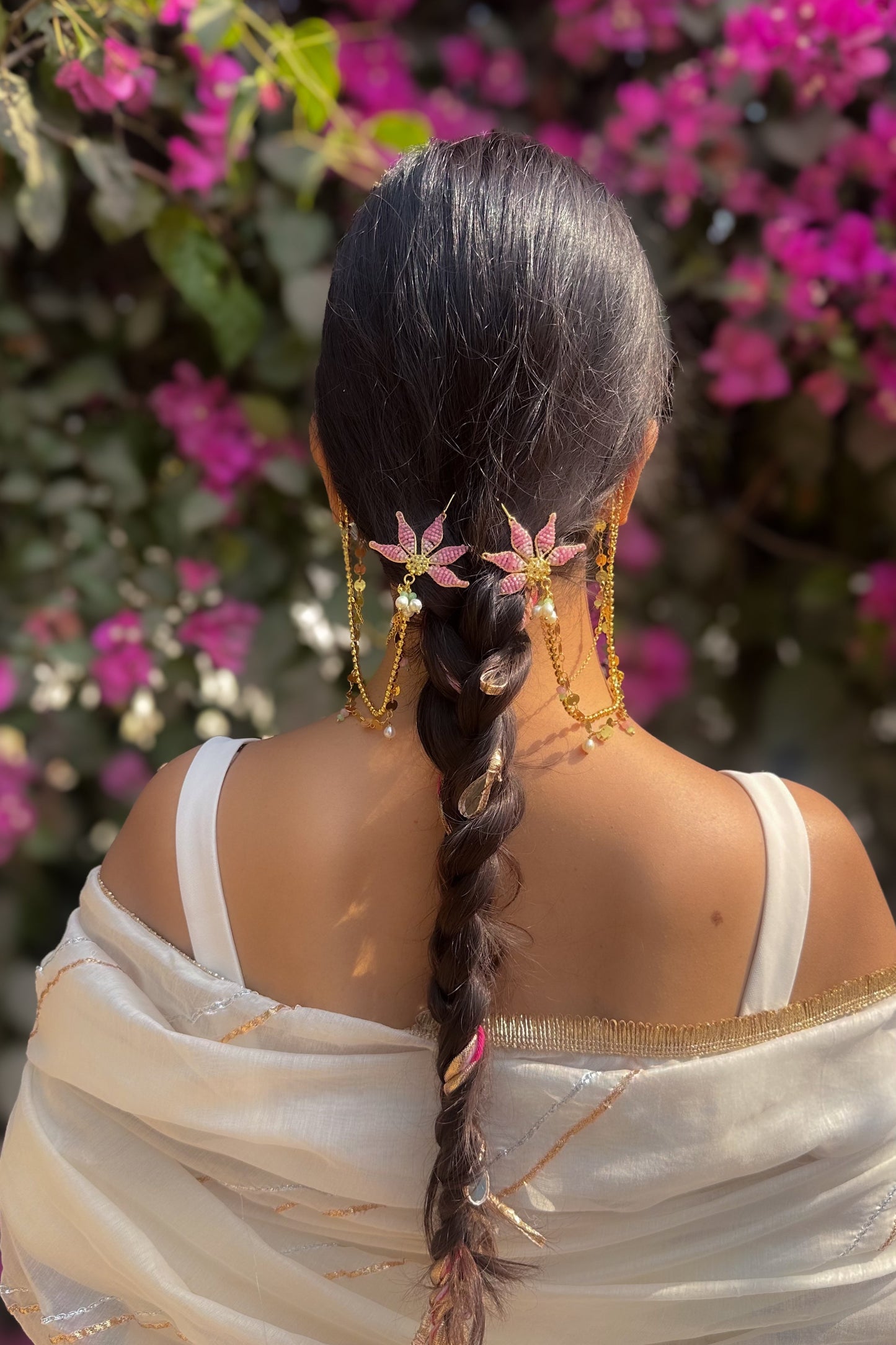 Woman with braided hair and floral decorations, wearing a white saree, against a pink flower background.
