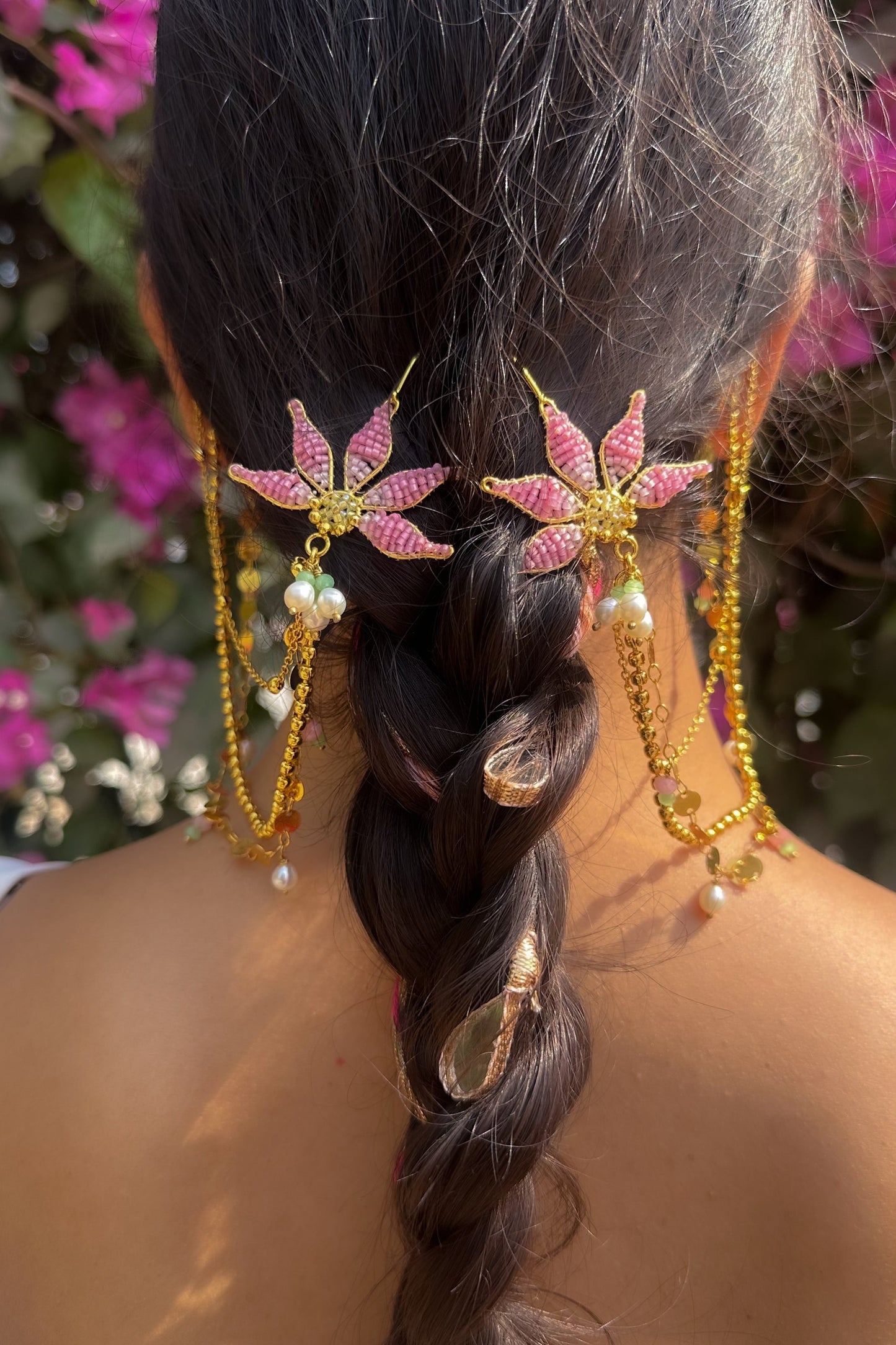 Decorative hair accessories with pink leaves and gold chains on a braided hairstyle against a blurred floral background.