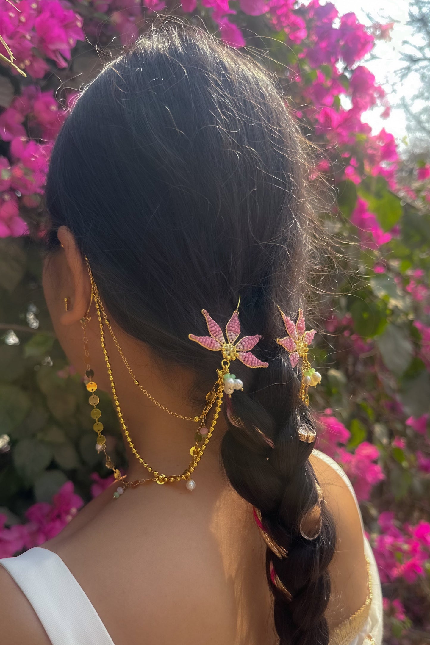 Person with braided hair wearing floral hairpins and gold jewelry against a pink flowering background