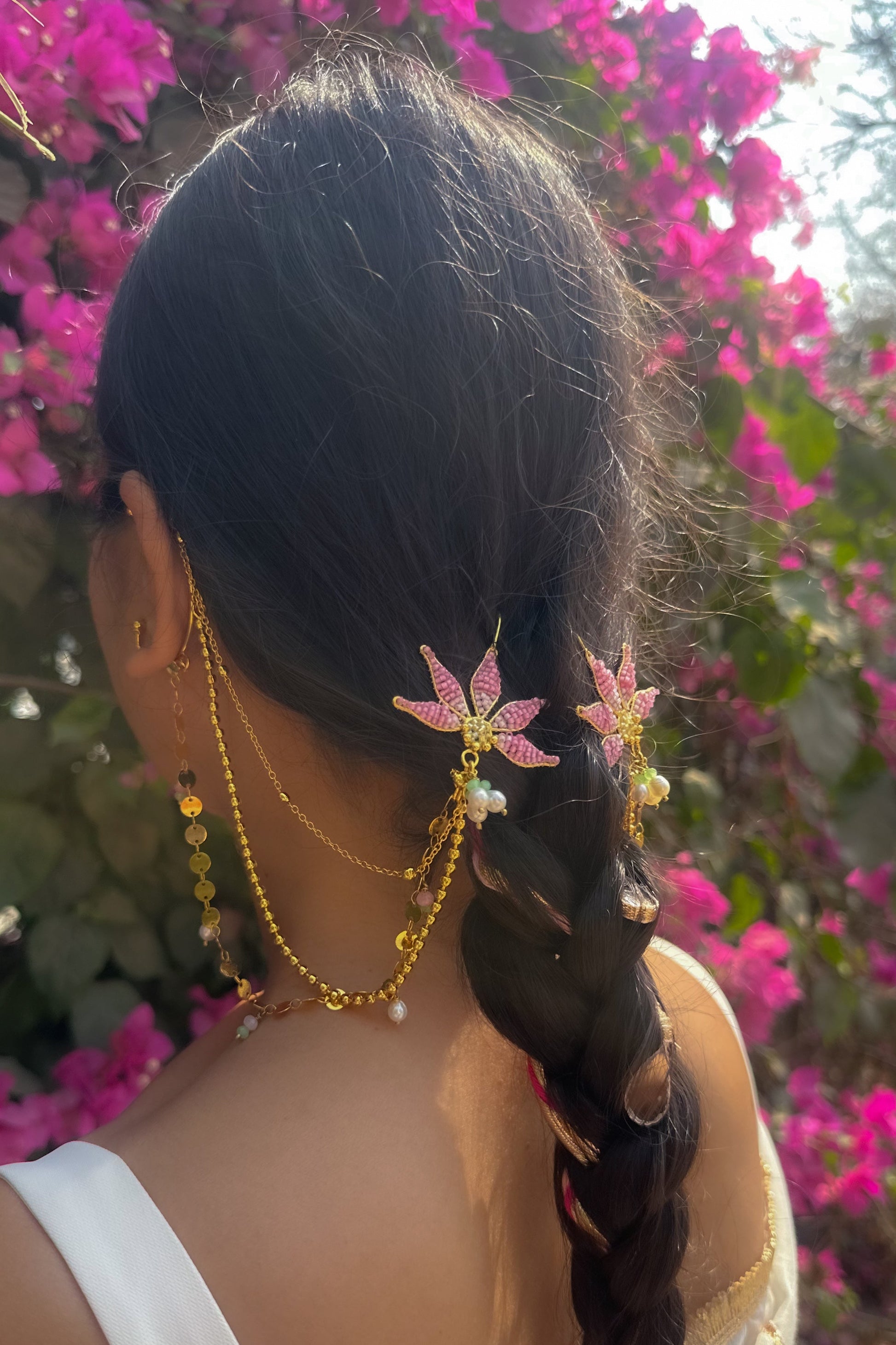 Person with braided hair wearing floral hairpins and gold jewelry against a pink flowering background