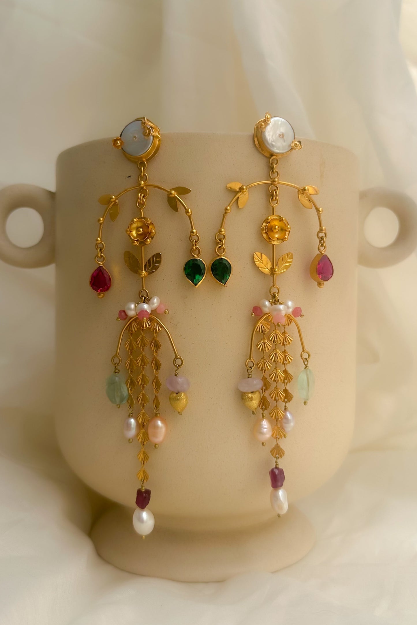 A pair of ornate gold-toned dangle earrings with multicolored gemstone accents and pearl-like drops, displayed on a beige ceramic cup against a soft white fabric backdrop.