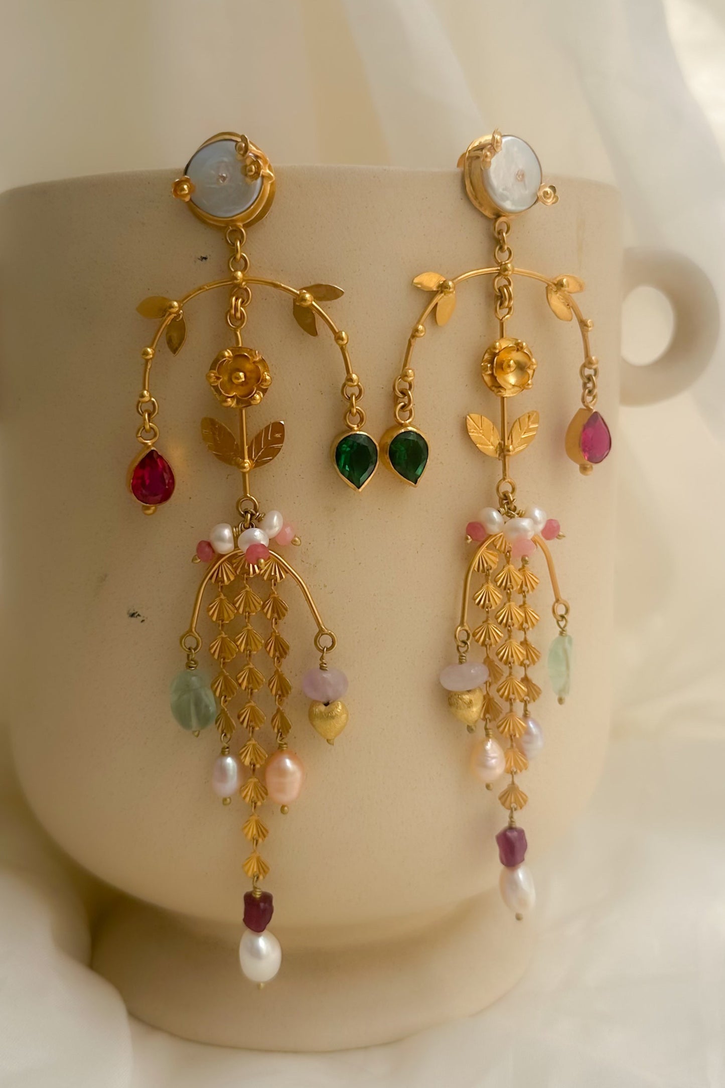 A pair of ornate gold dangle earrings with green and pink gemstone accents and white pearl-like beads, displayed on a beige ceramic cup against a draped white fabric background.