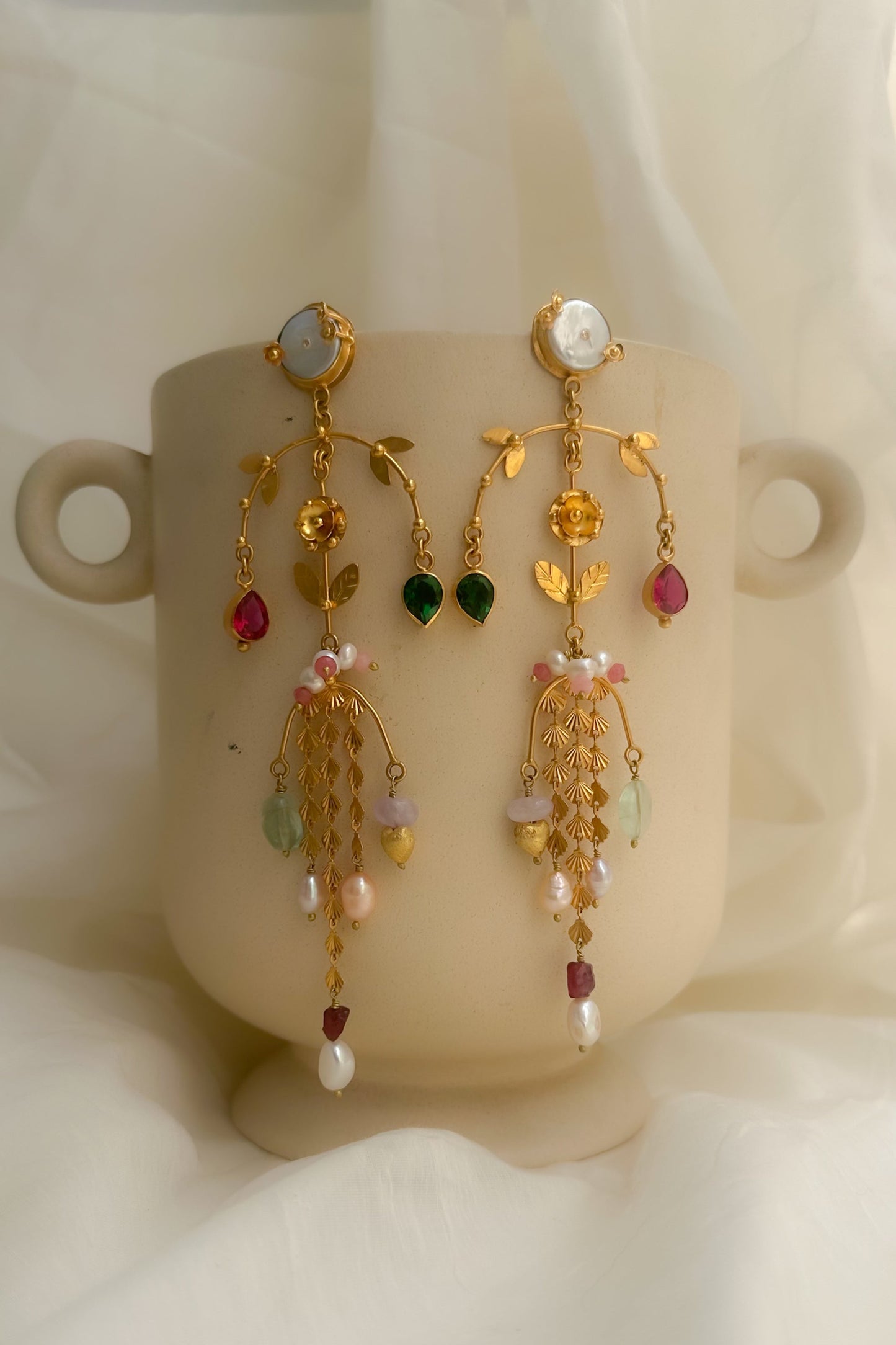 A pair of ornate gold dangle earrings with colorful gemstone accents, displayed on a beige ceramic cup against a soft white draped fabric background.