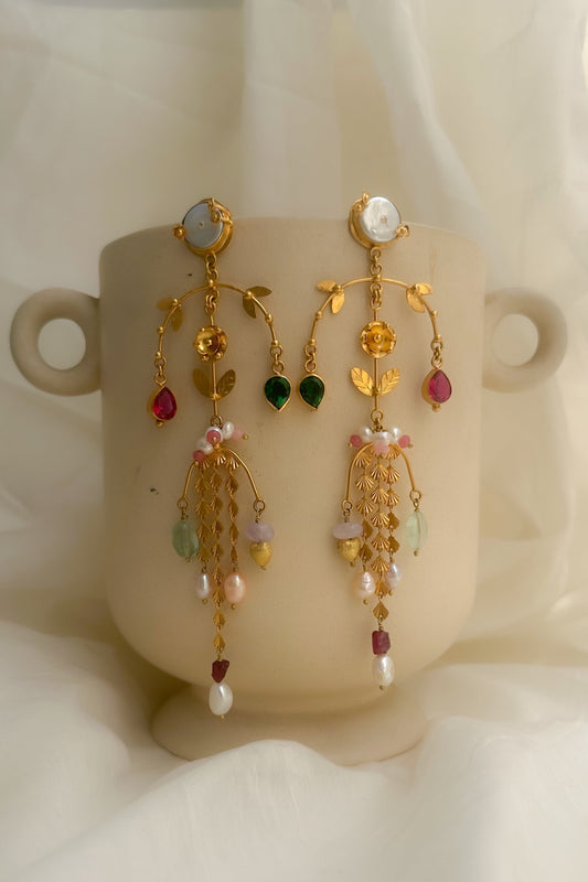 A pair of ornate gold dangle earrings with colorful gemstone accents, displayed on a beige ceramic cup against a soft white draped fabric background.