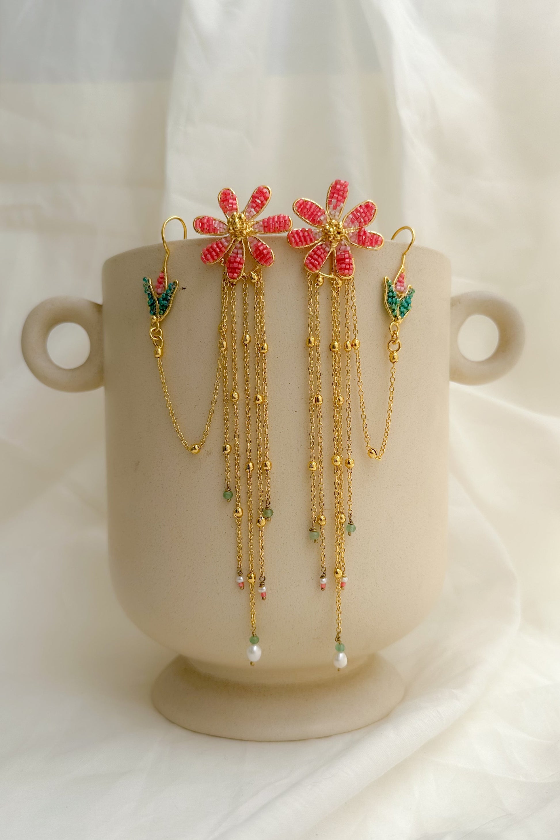 A pair of gold-toned dangle earrings with red flower motifs and green leaf accents, displayed on a beige ceramic cup against a soft white fabric backdrop.