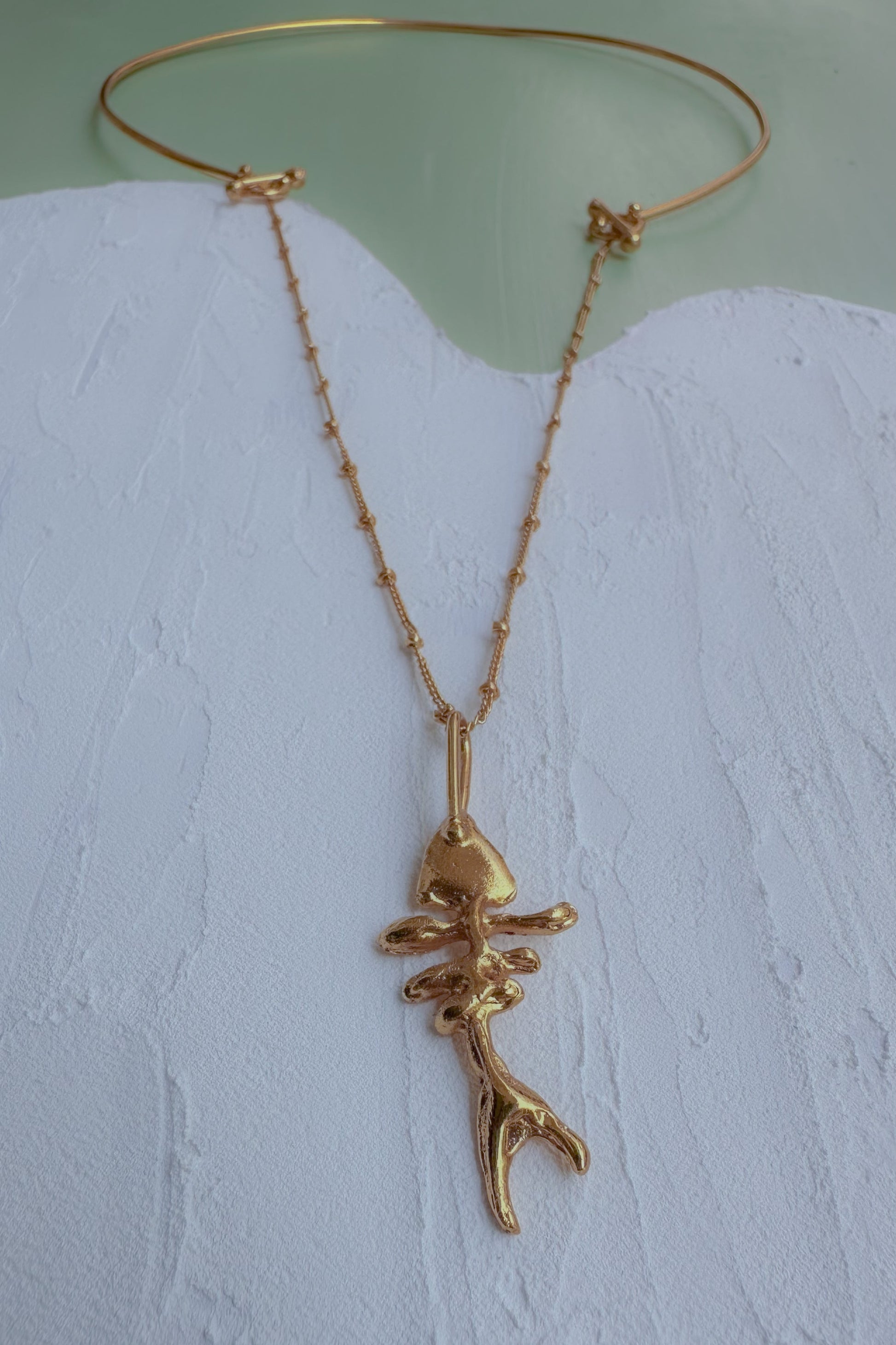 A gold-toned necklace with a delicate chain and a small abstract pendant, displayed on a textured white surface against a pale green background.