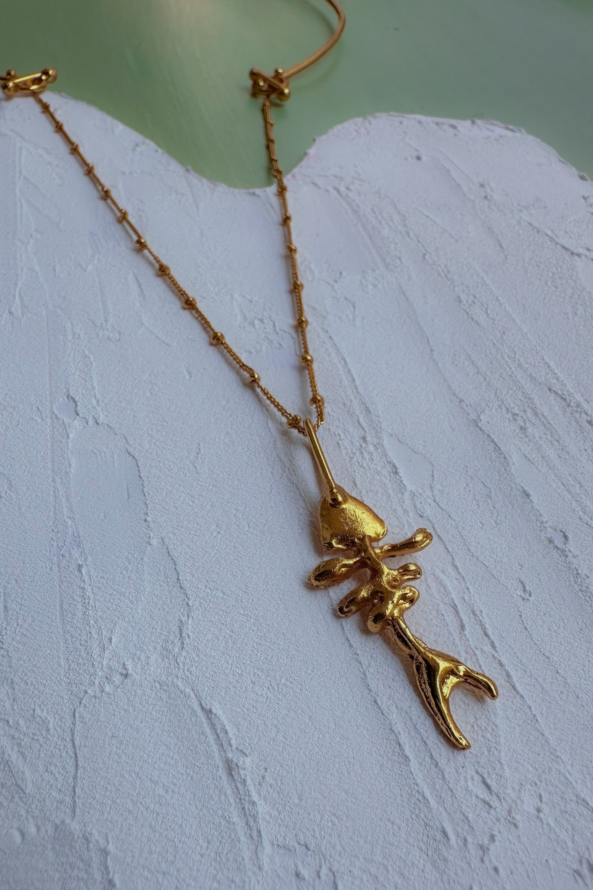 A gold chain necklace featuring a unique pendant shaped like a stylized, textured fish or sea creature, displayed on a white textured surface.