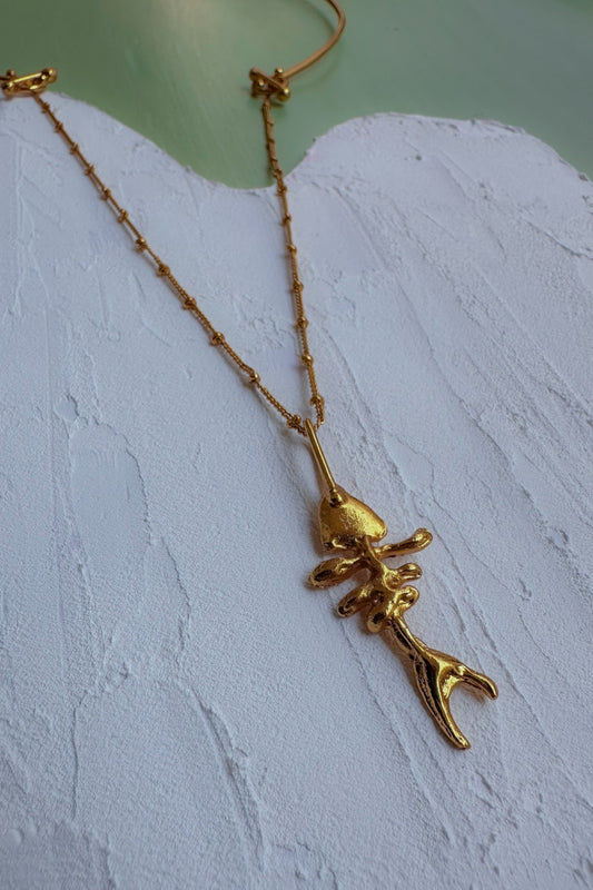 A gold chain necklace featuring a unique pendant shaped like a stylized, textured fish or sea creature, displayed on a white textured surface.