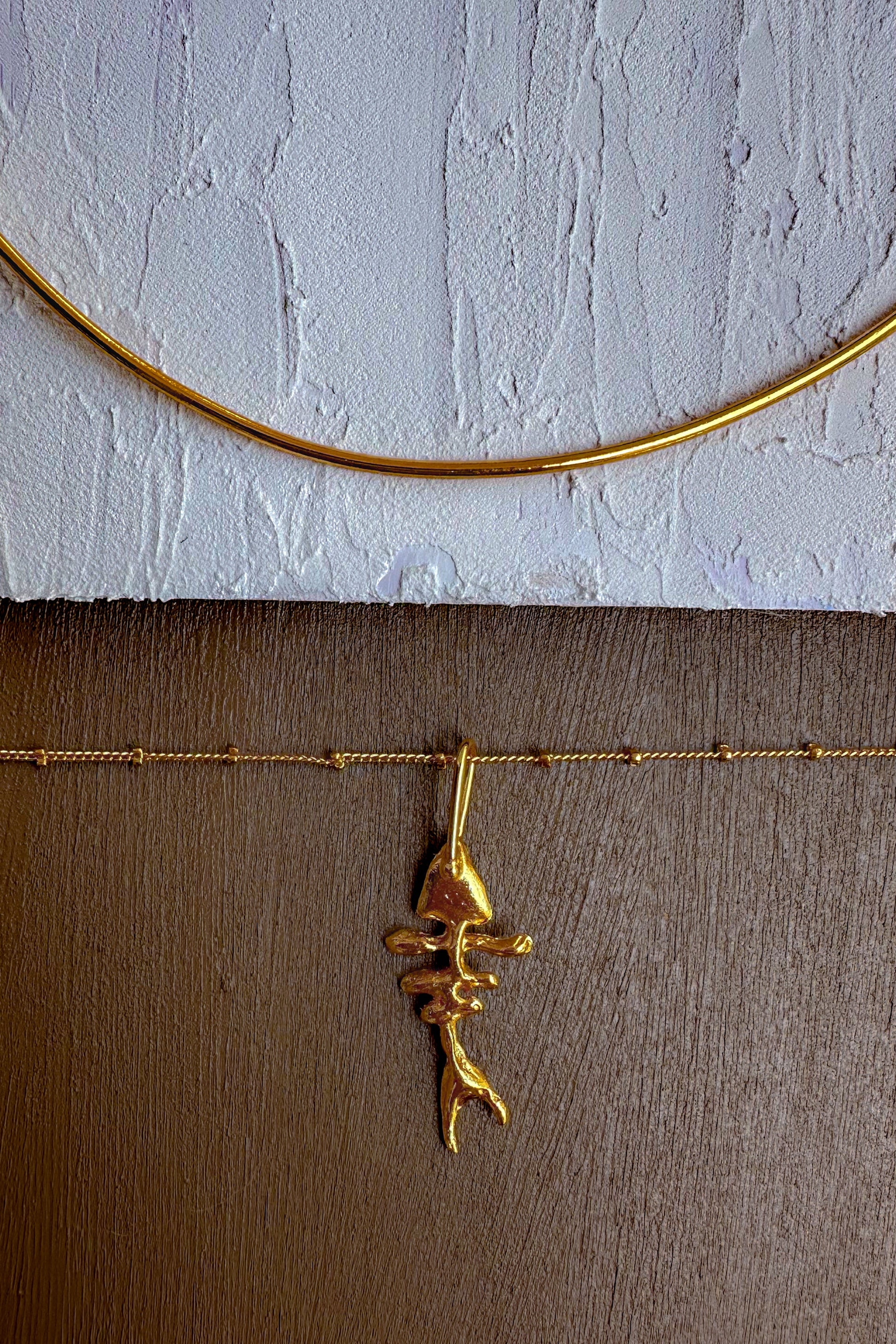 A gold necklace with a textured, organic-shaped pendant is displayed against a split background of a white textured wall and a brown wooden surface.