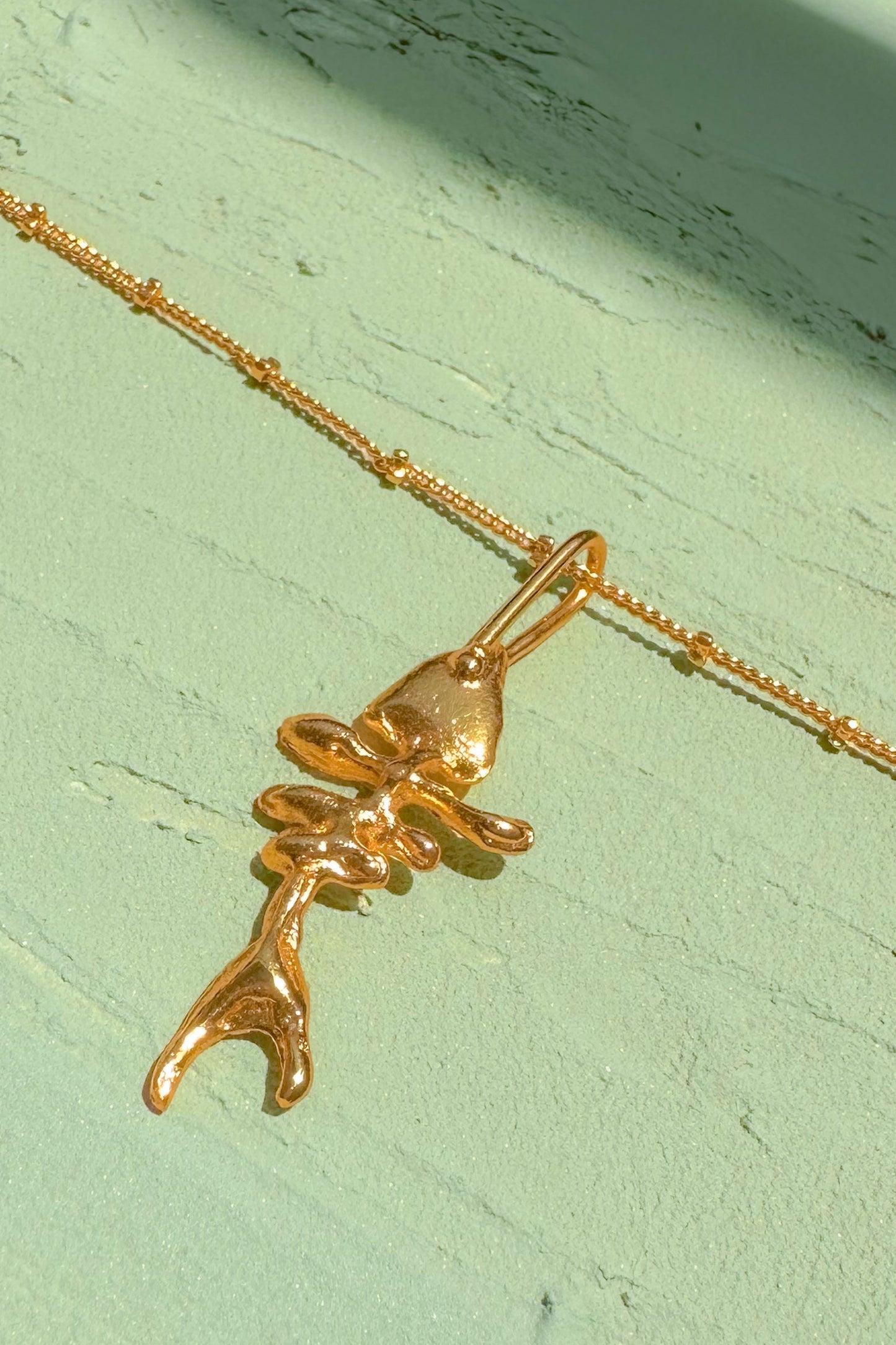 A gold pendant necklace featuring a stylized, abstract figure design, resting on a textured light green surface.