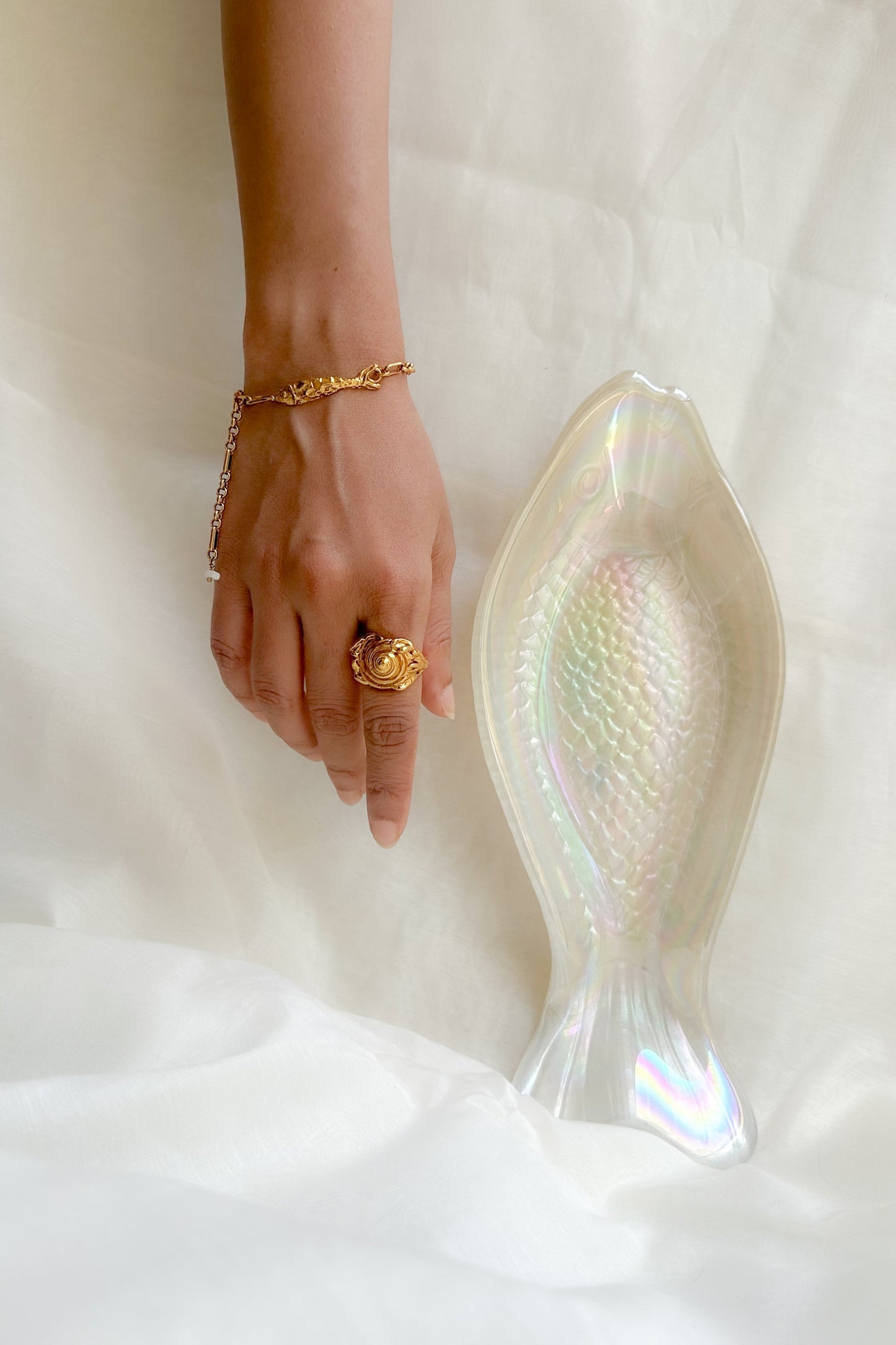 A close-up of a person's hand wearing a gold ring and bracelet, resting beside a translucent iridescent fish-shaped dish on a white fabric background.